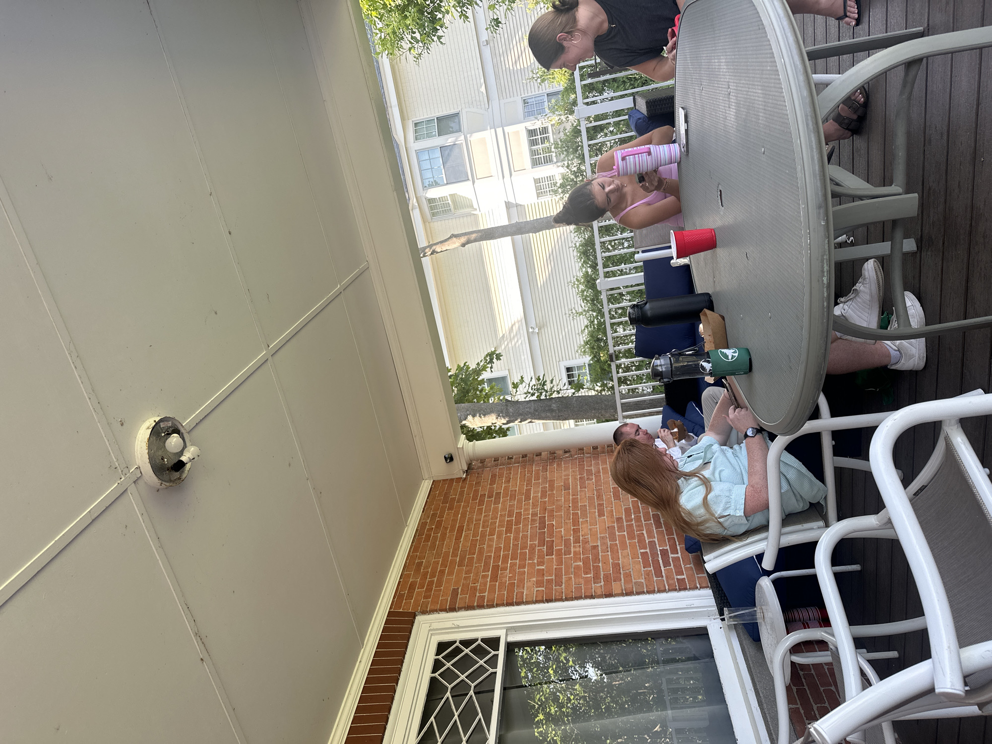 Four individuals are seated at a grey, oval outdoor table on a wooden deck, likely a porch or patio. The scene appears to be in Harbor Springs, USA, with a residential building visible in the background. One woman with long red hair, wearing a light blue striped short-sleeved shirt, is sitting at the table, looking down at her phone. Beside her, a man wearing a white shirt is also seated at the table, looking down. To the right, two other women are seated. One is wearing a pink tank top and looking at her phone, while the other, wearing a black t-shirt, is facing her. On the table, there are several items, including water bottles, a red cup, and what appears to be a small container of food. The porch ceiling has a visible light fixture, and brickwork is present on the left side of the image, suggesting the architecture of the building. The overall atmosphere is casual, possibly a gathering or meal.