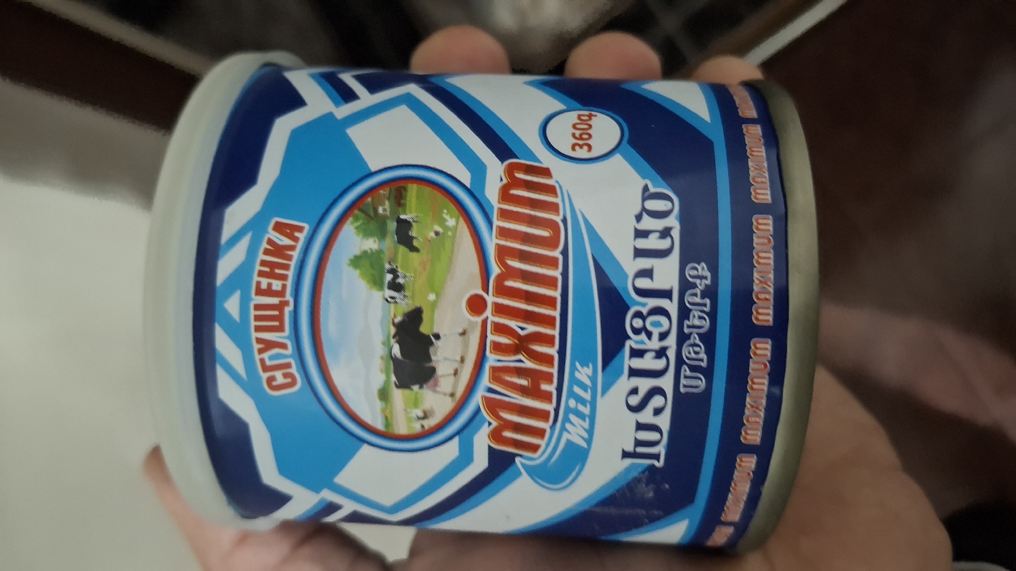 The image displays a person's hand holding a canned food product, likely indoors. The can is adorned with a vibrant blue and white label featuring geometric patterns. The primary brand name "MAXIMUM" is prominently displayed in red and gold lettering, with the word "MILK" positioned beneath it.
The label includes text in multiple languages: "СГУЩЕНКА" (Sgushchenka, meaning condensed milk) in red Cyrillic letters on the left side, and "ՄԱՔՍԻՄՈՒՄ" (Maximum) and "ԿԱԹ" (Kat, meaning Milk) in Armenian script on the right side. A small oval indicates the net weight as "360գ" (360 grams). Along the bottom edge of the label, the word "MAXIMUM" is repeatedly printed in a smaller font.
A circular emblem in the center of the label showcases a serene pastoral scene: several black and white cows are seen grazing in a lush green field under a bright blue sky, with trees in the background. The can is held at a slight angle, providing a clear view of its packaging design. The presence of Armenian text aligns with the provided context of Gyumri, Armenia. No specific time of day or weather details are discernible.