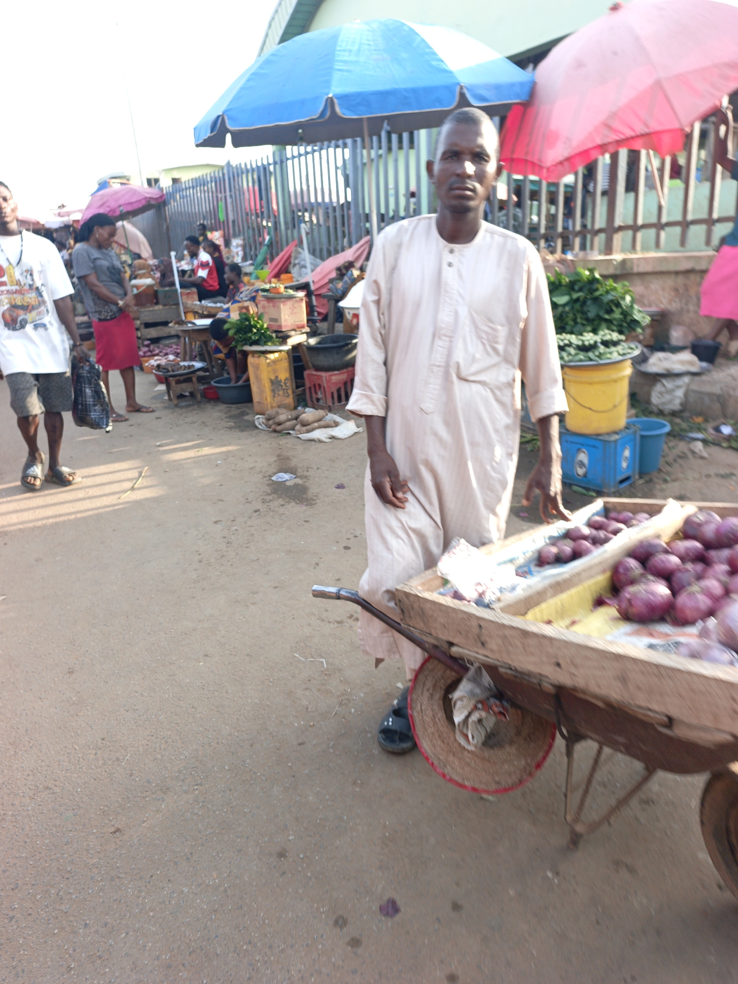 The image shows a street scene in Akwanga, Nigeria. In the foreground, a man in a light-colored robe stands next to a wooden wheelbarrow filled with red onions for sale. He looks directly at the camera. The wheelbarrow also has a hat hanging off the side. The road is paved. In the background, there are other people, some vendors under umbrellas. There are vegetables for sale, a blue plastic crate, and a yellow bucket. In the far background, there is a fence and a building.