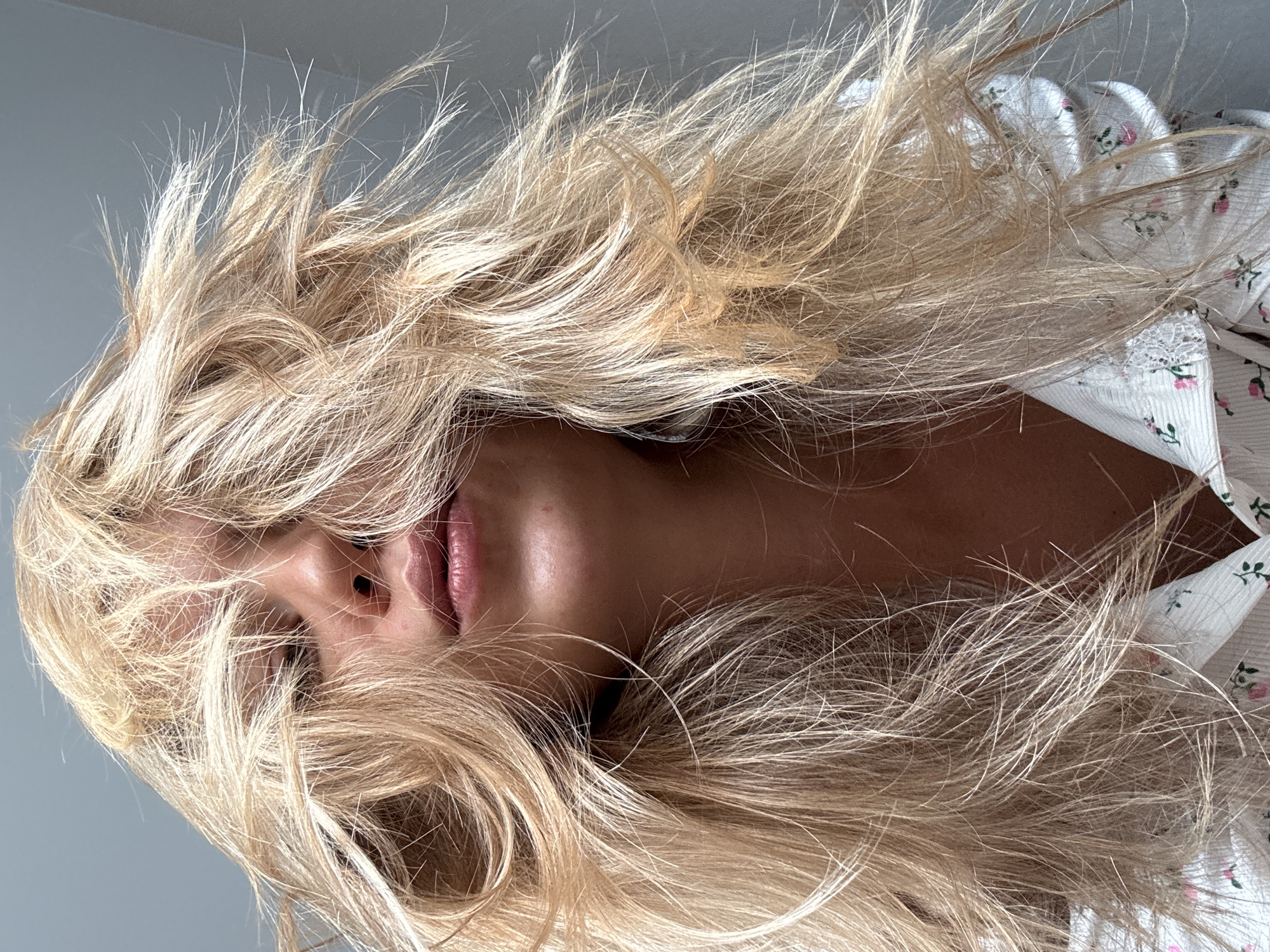 A person with long, blonde hair is seen from a low angle, with the hair covering much of the face. The person's lips and nose are visible through the hair. The person is wearing a white shirt with a floral pattern. The background is a plain, light grey color. The location is unknown.