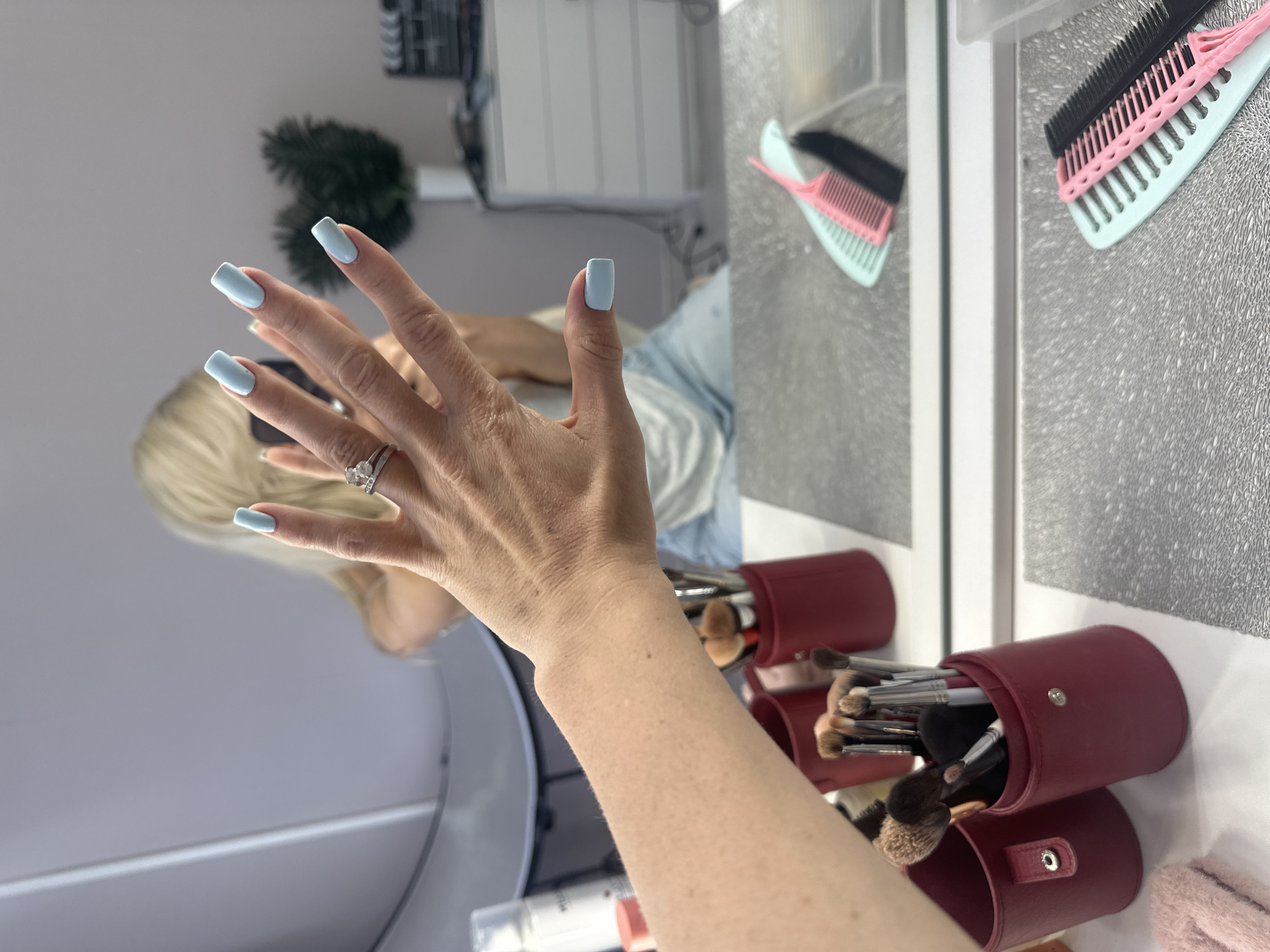 The image shows a person's hand with light blue acrylic nails, adorned with a diamond ring on the ring finger. The hand is positioned in front of a mirror, reflecting a blonde-haired individual's head and shoulders. In the background, on a reflective surface, are two burgundy makeup brush holders, one filled with various makeup brushes. To the right of the brush holders, a pink and black comb are visible. The setting appears to be a well-lit indoor space, possibly a vanity area or a room with a large mirror. The reflection of the person and the objects suggests that the photo is taken in a personal space, perhaps a home. The overall mood is neutral and focused on the presented elements. No specific time of day or weather is discernible. There is no visible text in the image.