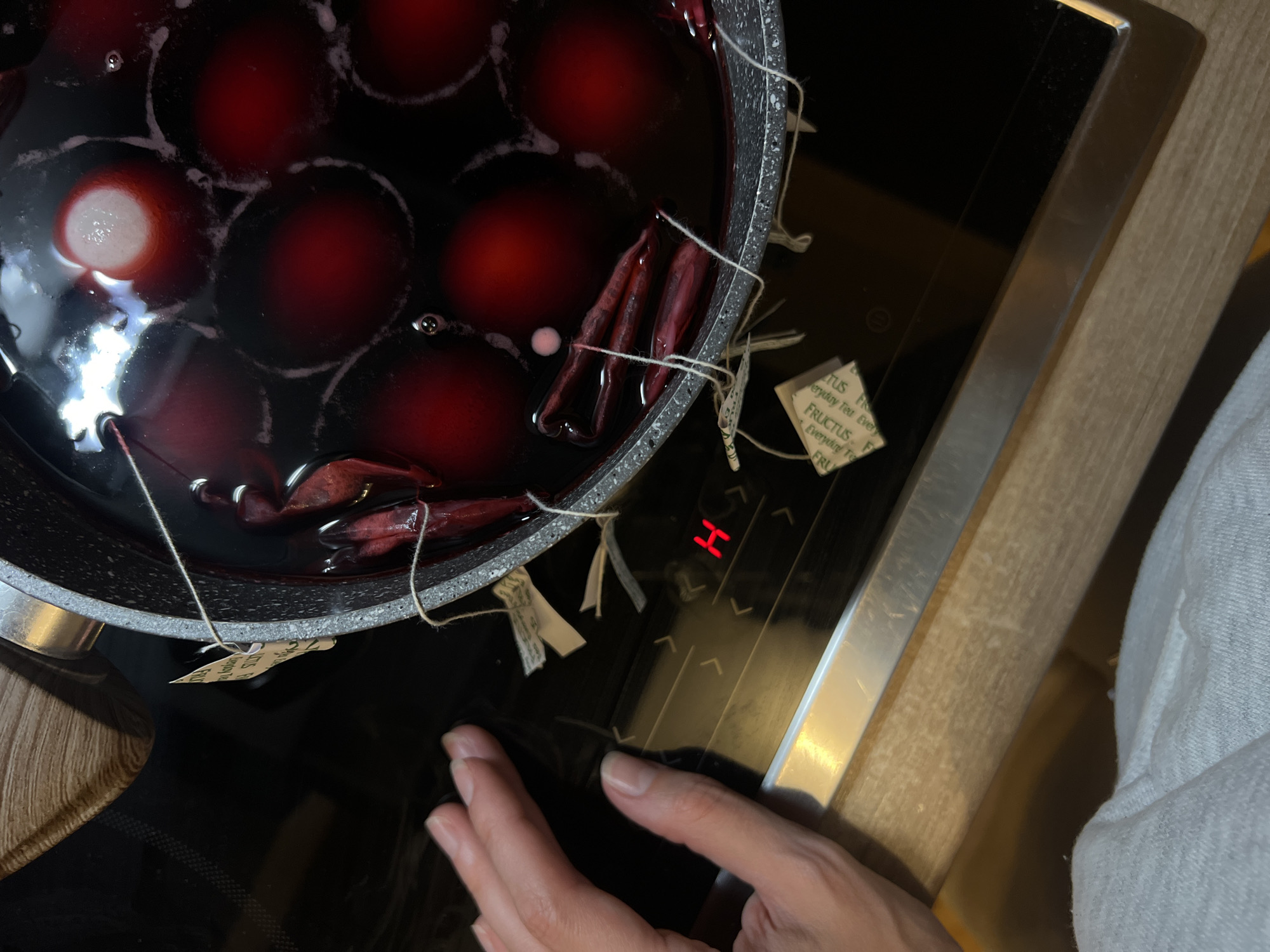 A top-down view captures a cooking scene, most likely in a kitchen, potentially in Bjeliši, Montenegro. A person's hand is visible near a modern stovetop. A pot sits on the stovetop, filled with a dark-colored liquid containing eggs being dyed. Several tea bags are steeping in the liquid, tied to the pot with string. The stovetop's digital display shows an "H," possibly indicating a hot surface. The surrounding environment includes elements of a countertop.
