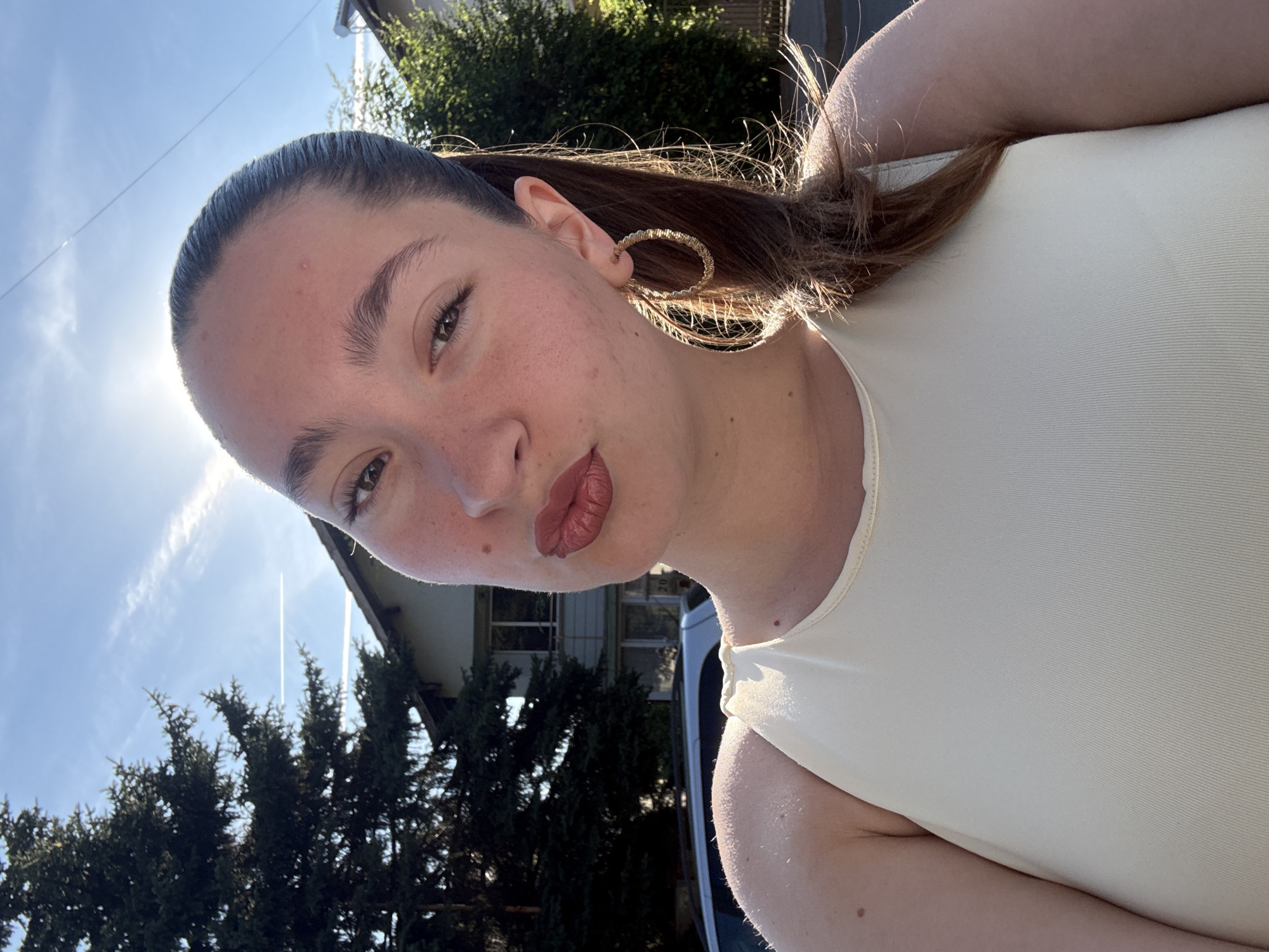 An outdoor, eye-level, medium shot shows a woman posing for a selfie in a suburban neighborhood. The woman is a fair-skinned young adult with dark hair pulled back in a ponytail. She is wearing a cream-colored tank top and large gold hoop earrings. She has on dark lipstick and is puckering her lips. The background shows a residential house with a large evergreen tree in front of it, and a white SUV parked in the driveway. The sky is bright blue with some wispy clouds. The sun is shining brightly from behind the woman's head, causing a lens flare. The location is identified as Sofia, Bulgaria.