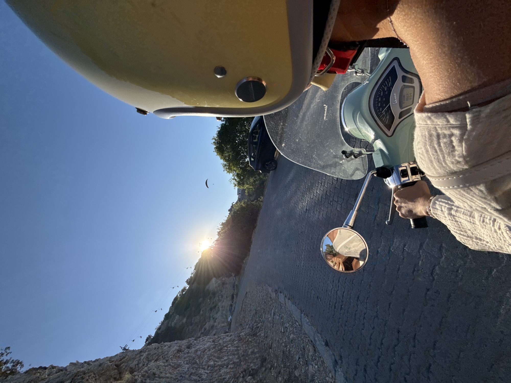 The image is a point-of-view shot from someone riding a light green Vespa scooter along a paved road that winds up a hillside. The rider is wearing a yellow helmet and a light-colored, textured shirt. The road is lined with a rocky, steep embankment on the left and vegetation on the right. The sun is setting, casting a warm glow and long shadows across the scene, and is positioned above the hillside, with rays extending outwards. In the background, a black car is parked on the side of the road, and a paraglider is visible in the sky. The scooter's dashboard is visible, displaying speed and other indicators. A mirror on the scooter reflects the rider's face and upper torso. The scene suggests a scenic drive in Fethiye, Turkey, possibly leading to a viewpoint.