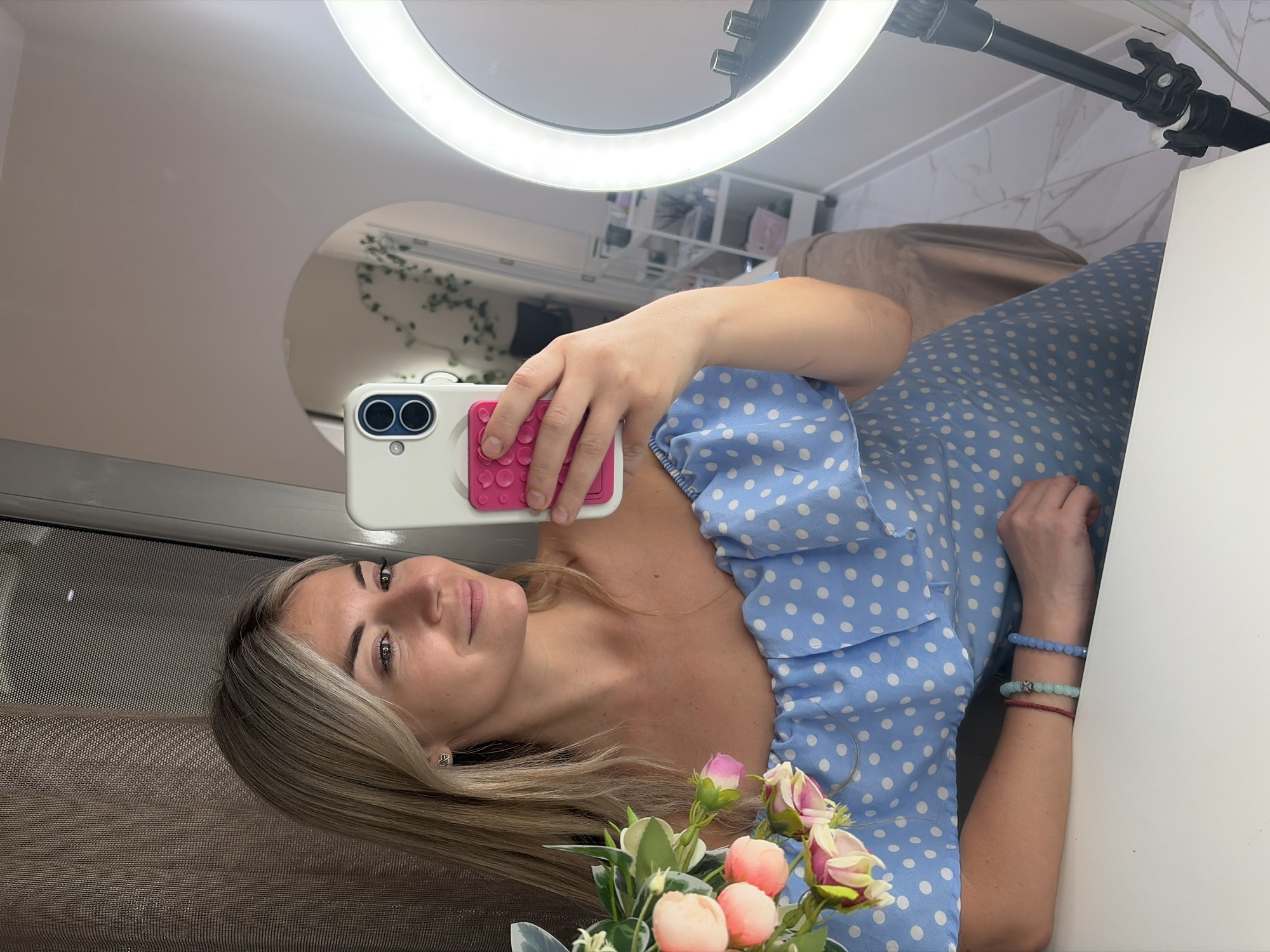 A blonde woman is taking a selfie in a room with light-colored walls and floor. She is wearing a light blue polka dot off-the-shoulder dress and several beaded bracelets on her left arm. She is holding a white smartphone with a pink phone case in her right hand. To the right of the woman, a ring light on a black tripod is visible. In the background, a white shelving unit is present, and a brown massage table is partially visible. The room appears to be a beauty salon or spa, possibly located in Budva, Montenegro, given the context.