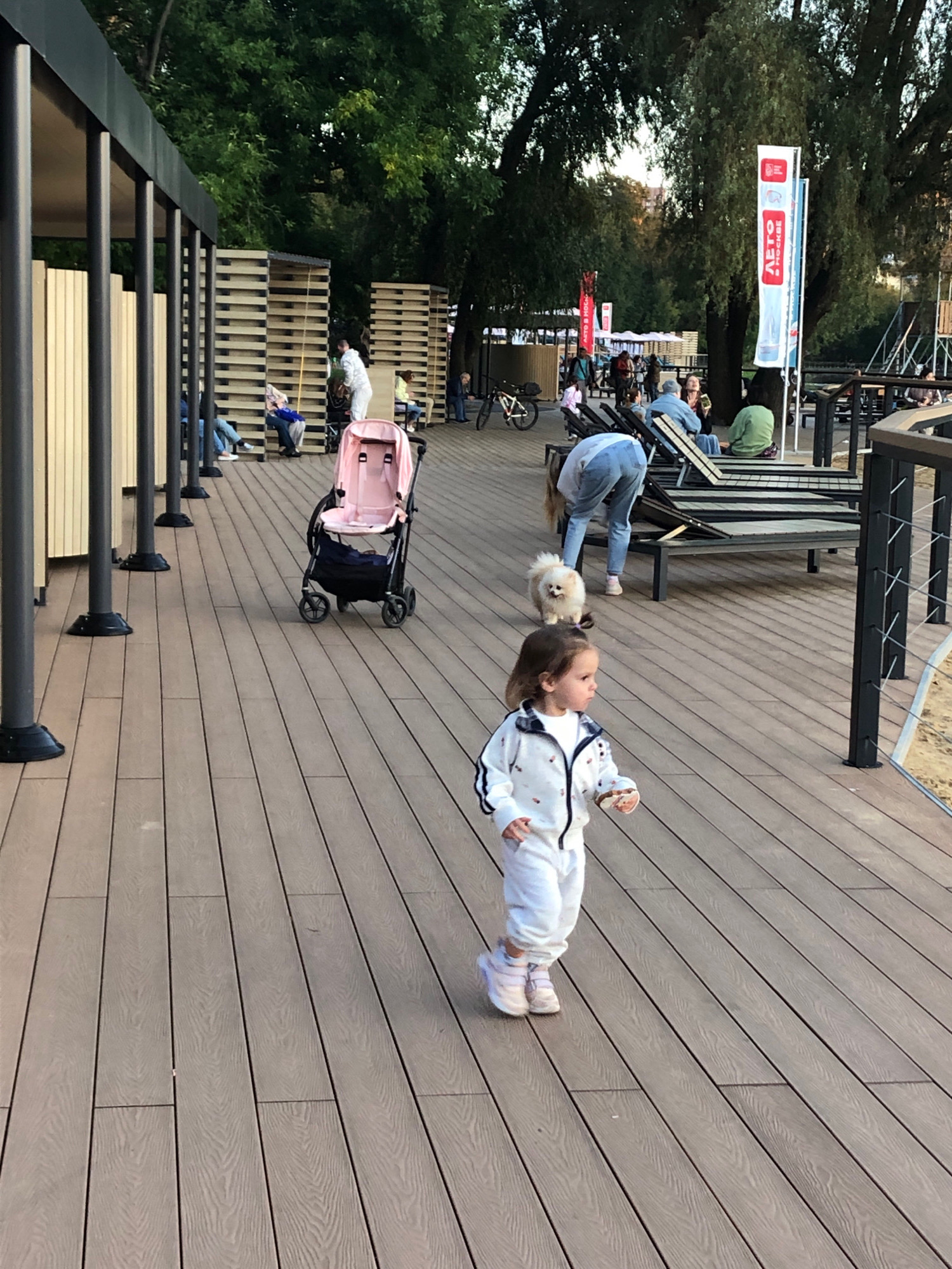 The image depicts an outdoor scene on a wooden boardwalk, likely in the city of Khimki, Russia, given the context of the flags with Russian text. In the foreground, a young child in a white tracksuit and pink sneakers is walking towards the right, holding a small object. Behind the child, a small fluffy white dog is walking. Further back, a woman in a white top and blue jeans is bending down near the dog. To the left, a pink baby stroller stands on the boardwalk. In the background, several people are seated on lounge chairs. There are also trees and modern architectural structures with vertical slats. Flags with Cyrillic text are visible in the distance, suggesting a public event or venue. The ground is made of brown wooden planks.