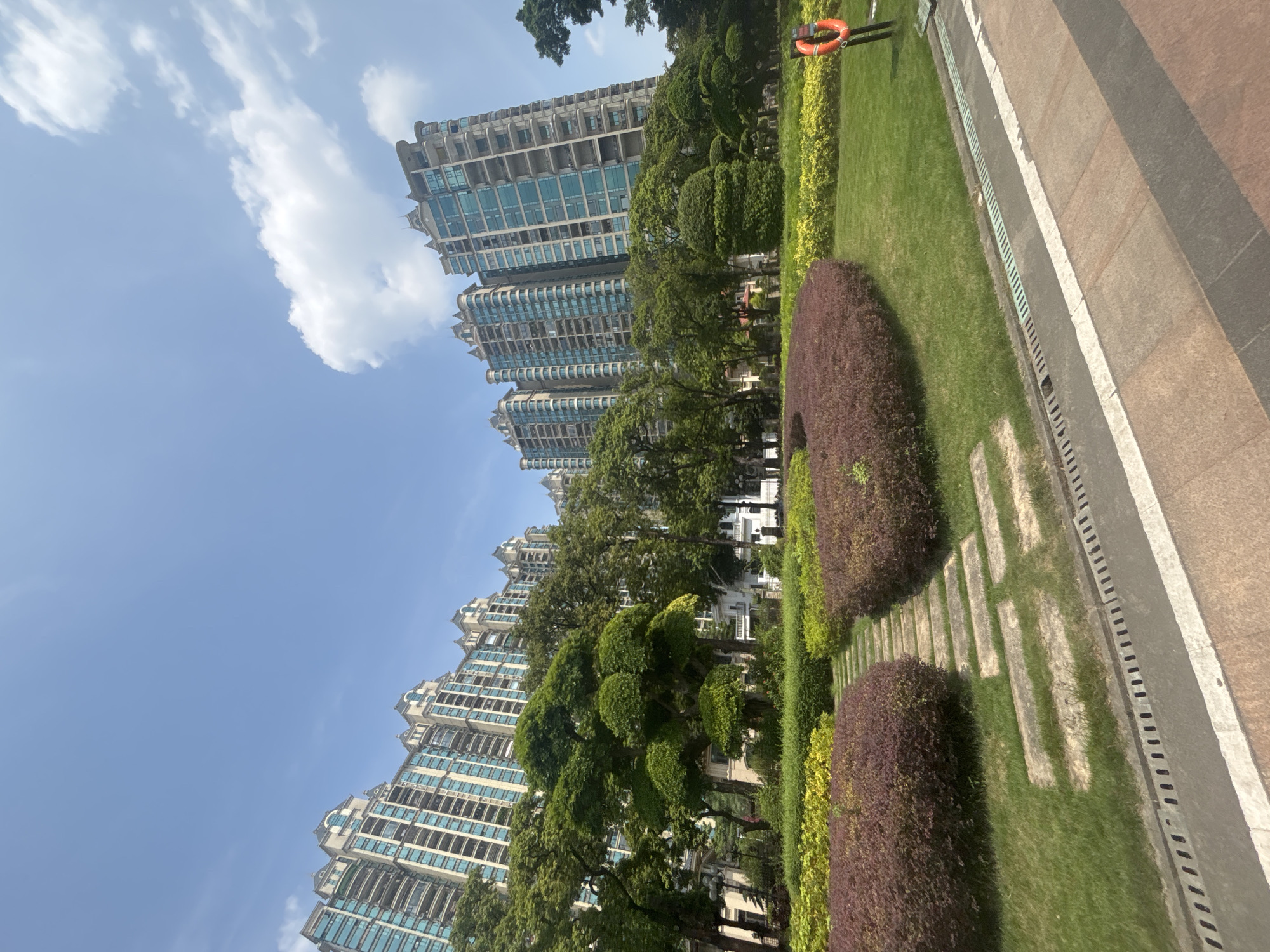 In the Nanhai District of China, a park scene unfolds beneath a clear blue sky dotted with white clouds. Tall, modern apartment buildings with light-colored facades and blue-tinted windows rise in the background, their multi-story structures characterized by balconies and decorative rooflines. In the foreground, a meticulously landscaped park features lush green lawns, vibrant red and yellow bushes, and a winding stone pathway. Mature trees, some with carefully sculpted canopies, provide shade and visual interest. A bright orange life preserver is mounted on a post in the middle-right of the frame, adjacent to a drainage grate running along the edge of a paved walkway. The overall impression is one of urban residential development integrated with green space.