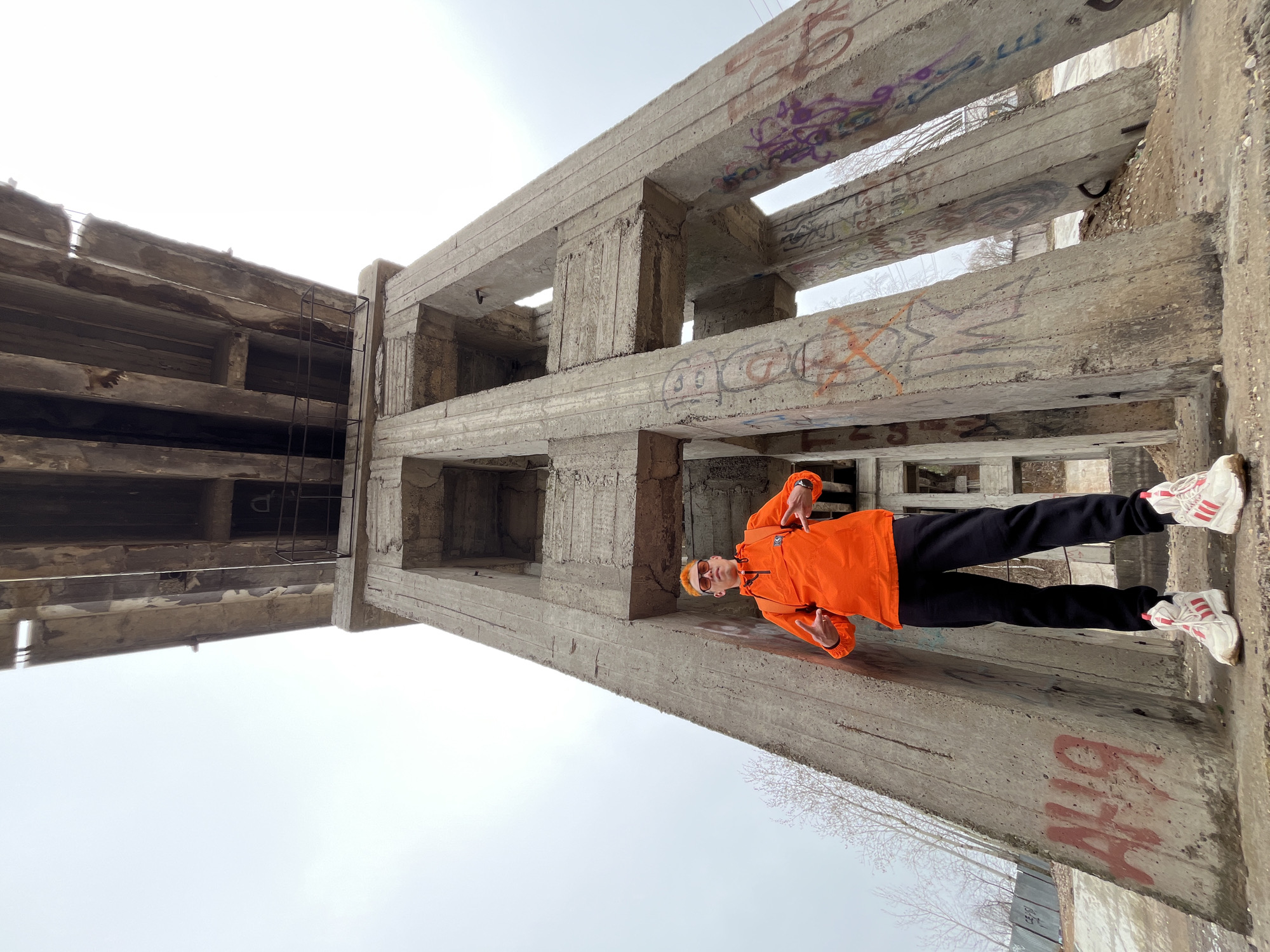 An individual stands on a large, weathered concrete beam, part of an extensive, possibly abandoned, concrete structure. The image, taken outdoors on an overcast day, shows a male with vibrant orange hair, wearing an orange anorak jacket, black pants, and white sneakers with red stripes. He is also wearing sunglasses and a small dark bag across his chest. He poses for the camera, making a peace sign with his left hand and pointing with his right.
The concrete structure features multiple massive beams and columns, displaying significant signs of age and extensive graffiti. The graffiti includes tags in various colors such as purple, orange, and red spray paint. Visible text and symbols on the concrete beams include "SOS", "BAD BOY", and a "DISCORD" logo. Partially visible red graffiti on a lower beam appears to include the letters "CHA" or "GHA". A metal ladder or grate is integrated into one of the vertical concrete sections on the left. Leafless trees are visible in the background, suggesting a late autumn or winter season. The sky is bright and overcast. The setting evokes an abandoned industrial or urban exploration (urbex) site in Chaikovsky, Russia.