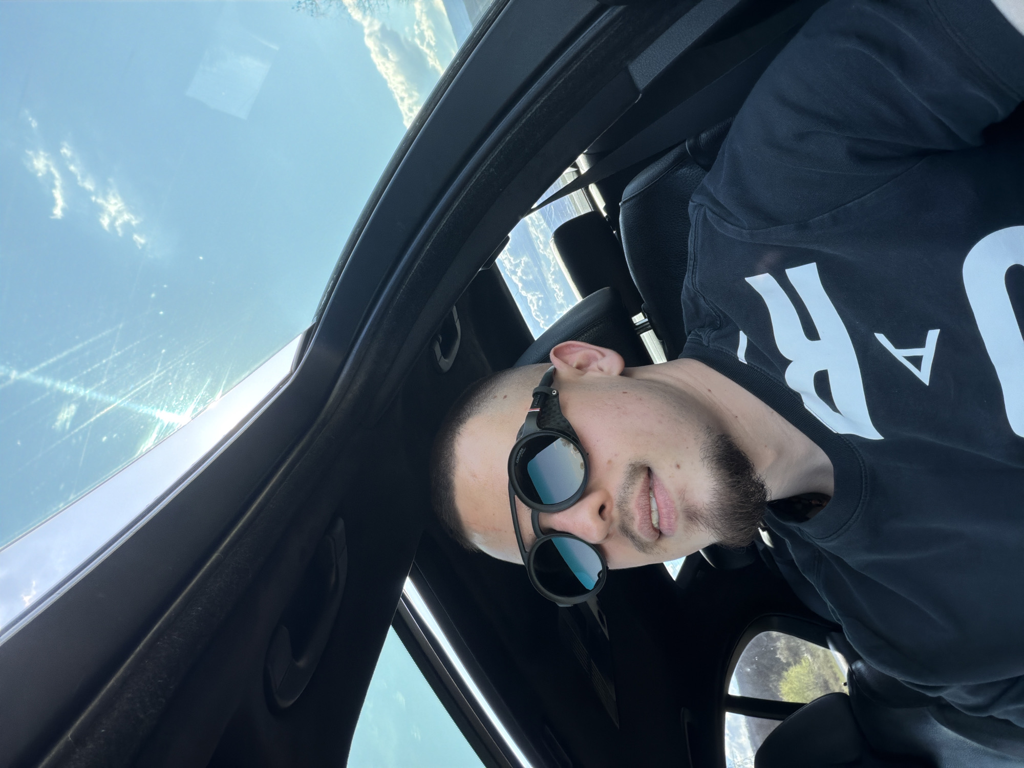 A medium shot of a light-skinned man with a short haircut and beard is taken inside a car looking upward through a sunroof. He is wearing sunglasses and a black t-shirt with large white lettering. The sunroof reveals a blue sky with sparse clouds. The car interior is black. The location cannot be determined to be Biaga, Bulgaria, from the image.