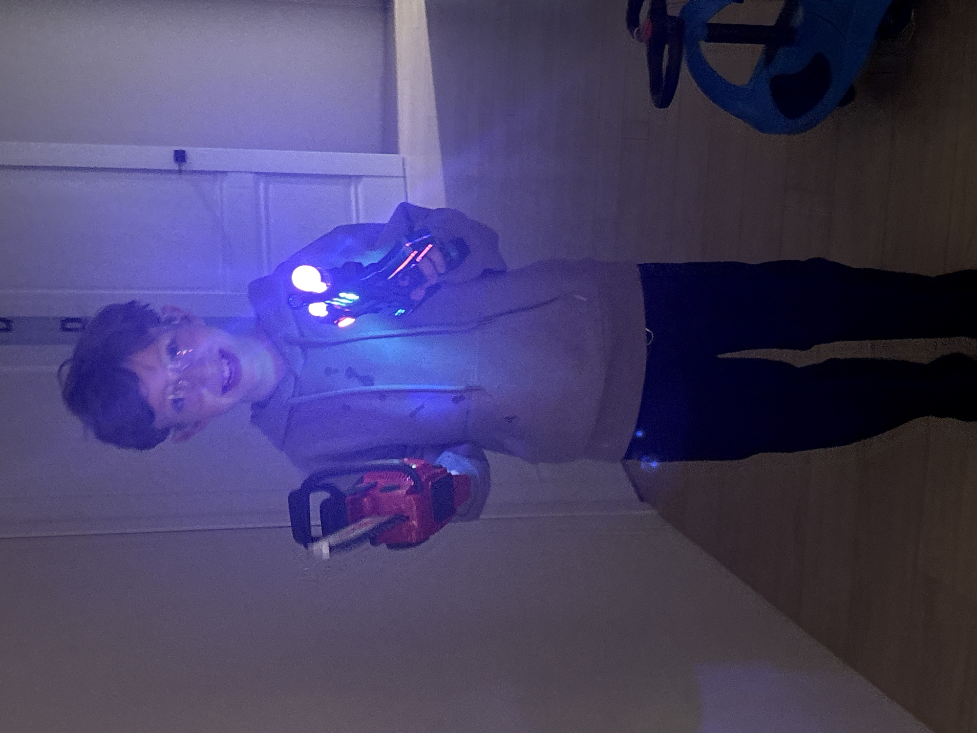 The image displays a young boy, appearing to be in his late childhood, standing in an indoor setting, likely a room within a home located in Chaikovsky, Russia. The boy is smiling broadly, indicating a playful and happy mood. He has short, dark hair and wears glasses.
He is holding two light-up toy blasters or futuristic guns, one in each hand. These toys emit bright, colorful LED lights, primarily blue, but also featuring red and green accents. The intense blue glow from the toys illuminates his face, hands, and the front of his light-colored hooded sweatshirt, creating a vibrant effect against the dim background. He is also dressed in dark trousers.
The room appears dimly lit, suggesting it is either evening, nighttime, or a space with low ambient light, which accentuates the luminosity of the toys. In the background, a light-colored door or closet is visible on the left side of the frame. To the upper right, part of a blue child's ride-on toy, possibly a PlasmaCar, can be seen. No visible text is present in the image.
