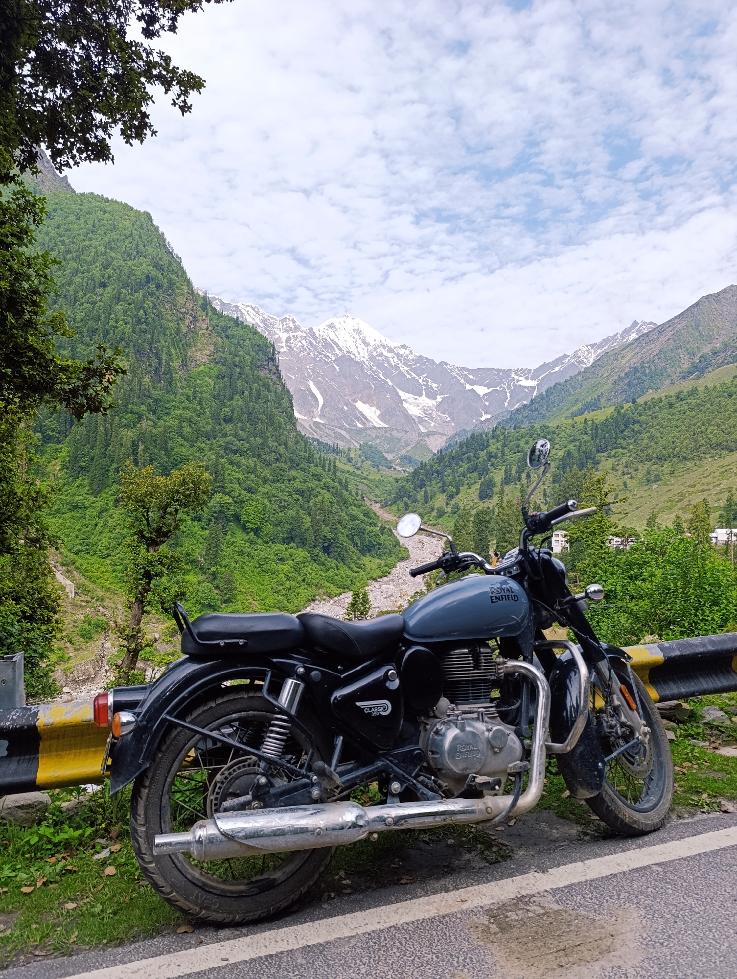 A grey Royal Enfield Classic 350 motorcycle is parked on the side of a paved road in Dhundi, India. The motorcycle is positioned at an angle, with its front wheel slightly turned towards the left. It is facing away from the viewer, with the rear of the bike closer to the camera. A yellow and black striped barrier runs along the edge of the road, behind the motorcycle.
The background is dominated by a lush green valley, with steep, tree-covered mountains on either side. In the distance, snow-capped peaks rise against a partly cloudy sky. The weather appears overcast, but the clouds are scattered, allowing some sunlight to illuminate the landscape. A riverbed is visible in the valley, with rocks and a small stream. The vegetation is dense, with a mix of trees and shrubs. The overall impression is one of a scenic, mountainous location, likely a popular spot for motorcycle riders. There are no people visible in the image.
