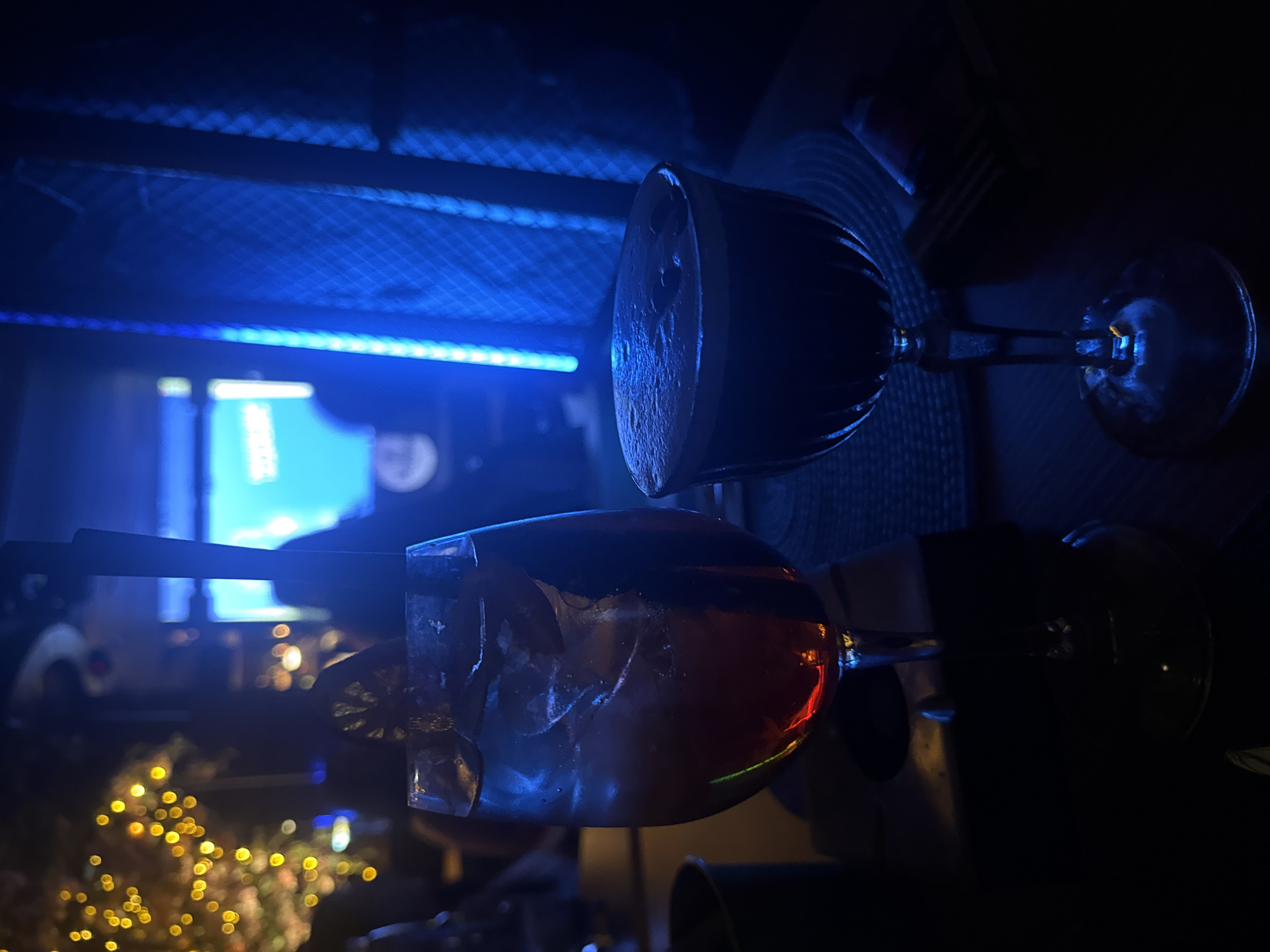 Two cocktails are visible in a dimly lit setting, likely a bar or restaurant. The cocktail on the left is in a stemmed glass and appears to be a reddish-amber color with ice and a garnish. The cocktail on the right is in a coupe glass and is dark in color with a foamy head. In the background, there are blurred lights, some of which are yellow and strung like fairy lights, and a bright blue neon light. The environment suggests a nightlife or entertainment venue. There are no discernible people clearly visible. This image was taken in Sofia, Bulgaria.