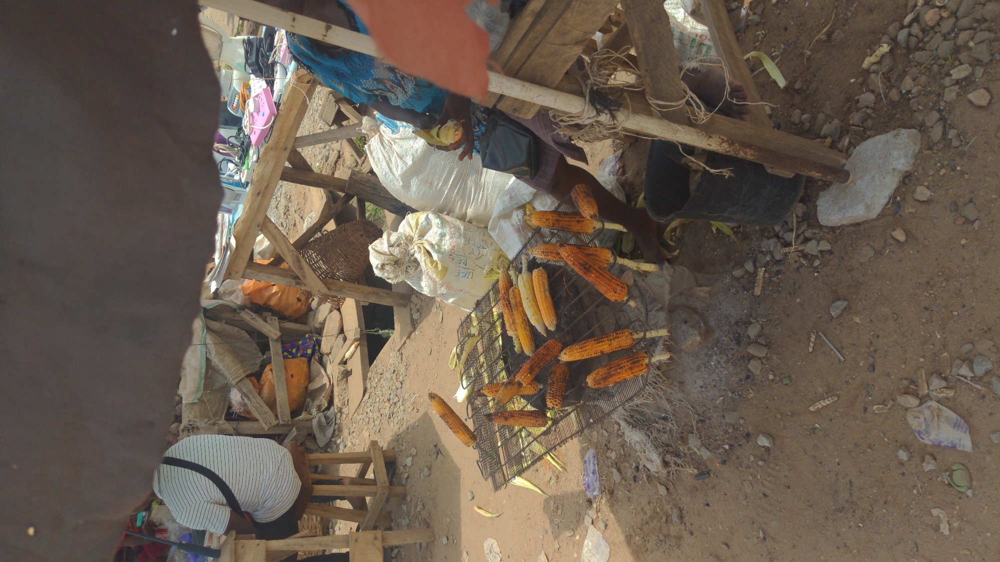 This outdoor scene captures an informal market setting, likely in Ado, Nigeria, with a primary focus on food preparation. The ground is bare earth, littered with small stones, dust, and scattered debris like plastic and paper.
In the center, a metal grill holds numerous corn cobs in various stages of roasting. Some cobs are light yellow, while others are a darker, charred orange-brown, many with sticks inserted for easy handling. Smoke or heat haze is visible around the grill, suggesting active use.
Two individuals are present. In the foreground, a person's back is partially visible, clad in a white and blue striped shirt and dark pants, leaning forward. Further back, another person, wearing a blue patterned outfit, is seated or crouched near the grill, with a hand holding a corn cob.
Around the grill, several large white sacks are visible, with one prominently displaying the word "RICE." Another sack has partially visible text including "PEF." A woven basket rests on a makeshift wooden stand, and a black bucket sits on the ground. The background features various wooden structures, likely stalls constructed from rough lumber, with glimpses of other market goods like small appliances or electronics on shelves.
The activity clearly depicts the roasting of corn for sale, indicating a street food vendor in action. The natural lighting suggests it is daytime. The scene conveys the busy, functional atmosphere of a local market.