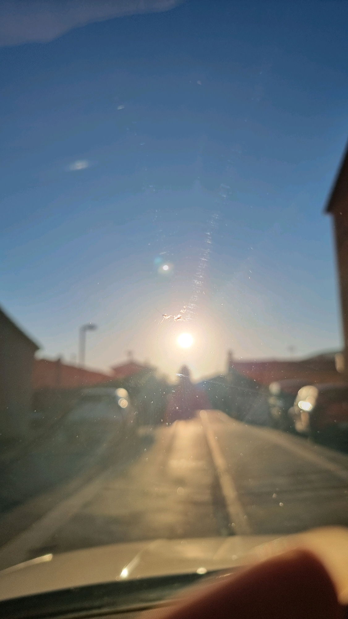 The image shows a view through a windshield, likely from inside a vehicle, looking towards a street lined with houses. The sun is low in the sky, creating a strong glare and lens flare, indicating it is either sunrise or sunset. The sky is a clear blue. The road ahead is paved, with a white line marking the center. Out-of-focus houses with reddish roofs are visible on either side of the street. A street lamp is visible in the distance on the left. The windshield has visible specks of dirt or water droplets. A blurred finger is visible in the foreground, partially obscuring the bottom right of the frame. While the image does not explicitly show specific landmarks of Sant Joan Les Fonts, Spain, the architectural style of the houses and the general environment are consistent with a residential area in a Spanish town or city.