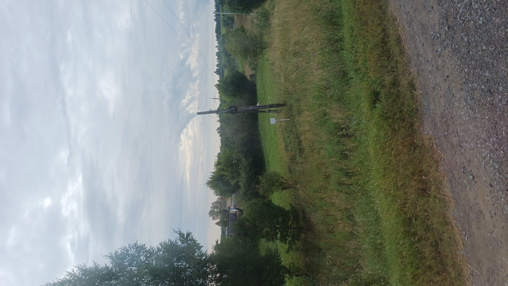 The image depicts a rural landscape under an overcast sky. In the foreground, a gravelly dirt road runs along the bottom edge. To the right of the road is a grassy area with tall, dry grass and some green vegetation. Further into the scene, there is a field of green grass with scattered bushes and trees.
Prominently featured in the mid-ground is a tall, weathered wooden pole with what appears to be a metal ring near the top. A small white sign is visible at the base of the pole. To the left of this pole, a cluster of houses with dark roofs is nestled amongst trees. A few more houses and trees are visible in the distance, suggesting a small settlement.
In the background, rolling hills covered in trees stretch to the horizon. Power lines can be seen extending across the upper right portion of the image. There is no visible human activity, and the scene appears to be peaceful and undeveloped. The environment suggests a location in the countryside of Buranovo, Russia, characterized by its natural beauty and traditional rural architecture.