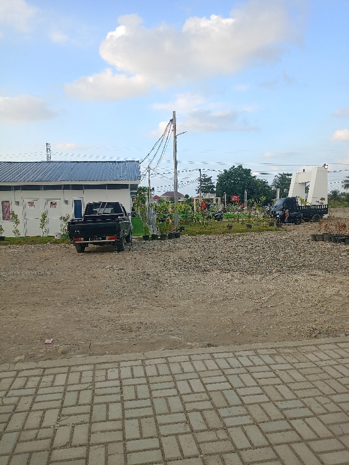 The image displays an outdoor scene in Meureudu, Indonesia, under a blue sky with scattered white cumulus clouds. The immediate foreground features a light grey paver block surface, arranged in a basket-weave pattern. Adjacent to this, the ground is unpaved, composed of dirt and gravel, with a small piece of pink litter visible on the left.
In the midground, a single-story white building with a dark blue corrugated metal roof is present on the left. It has multiple white doors and small rectangular windows. A black pickup truck, with a license plate "BL 8415" visible on its rear, is parked facing away from the viewer, positioned in front of the building. To the right of the building, a landscaped area contains plants in black plastic pots and a utility pole supporting multi-colored overhead wires. Further right within this area, a small black flatbed truck is parked, facing right, near a red and white sign structure and several parked motorcycles or scooters. A large, white, angular monument or archway is visible on the far right of the midground. The ground beyond the landscaped section is also unpaved and gravelly.
The background includes a faint metal lattice tower on the distant left and some distant buildings with reddish roofs partially obscured by trees. No individuals, discernible actions, or interactions are present in the scene.