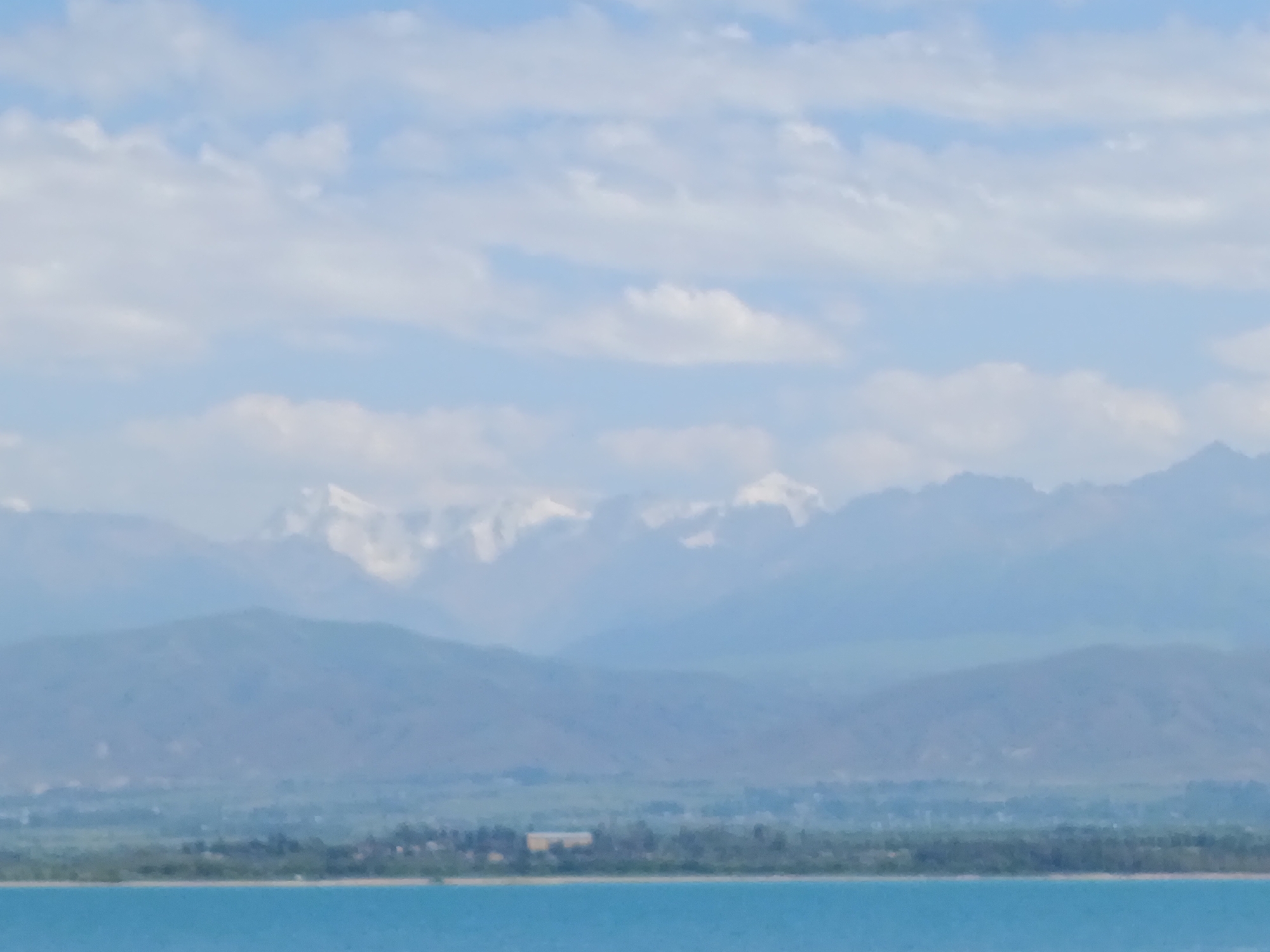 The image depicts a vast, serene landscape with a clear blue sky filled with scattered white clouds. In the foreground, a tranquil turquoise body of water stretches horizontally across the frame. Beyond the water, a verdant shoreline is visible, lined with scattered trees and what appears to be a small settlement or resort area. Further in the background, majestic snow-capped mountains rise, their peaks partially obscured by a light haze. The overall impression is one of natural beauty and peacefulness, likely in a location like Kyzyl-Suu, Kyrgyzstan, known for its stunning mountainous terrain and proximity to water bodies.