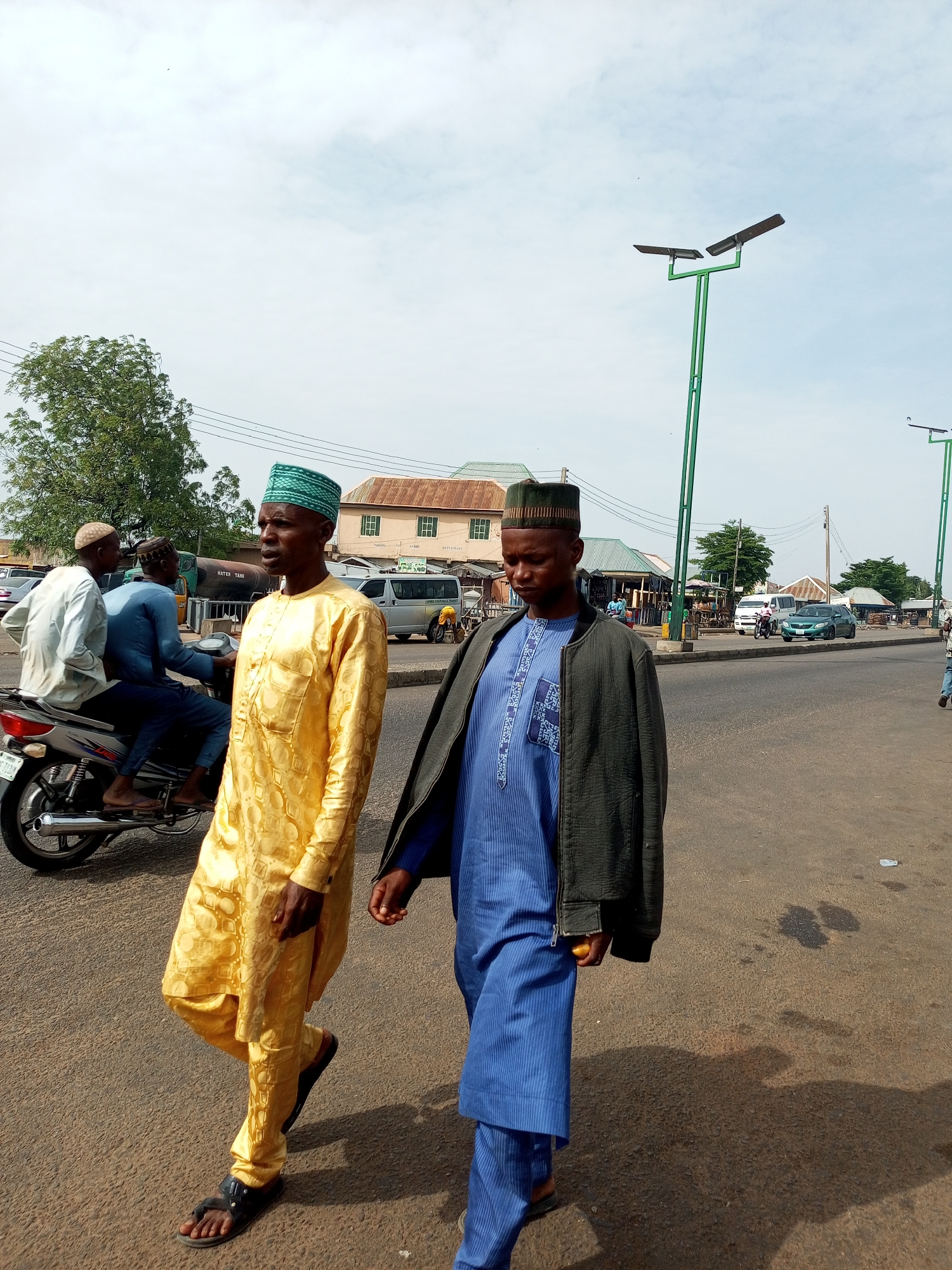 Two men in traditional Islamic attire walk on a paved road in Gombe, Nigeria. The man on the left wears a yellow patterned robe and cap, while the man on the right is dressed in a blue robe and dark jacket, also wearing a cap. In the background, several vehicles are visible, including a motorcycle with two passengers, a water tank truck, a white van, and a green car. Buildings with corrugated metal roofs line the street, and tall green lampposts with solar panels are strategically placed along the road. The sky is overcast, and the sun casts shadows on the ground. The scene depicts a typical urban streetscape in Gombe, likely a commercial area due to the presence of various shops and transportation.