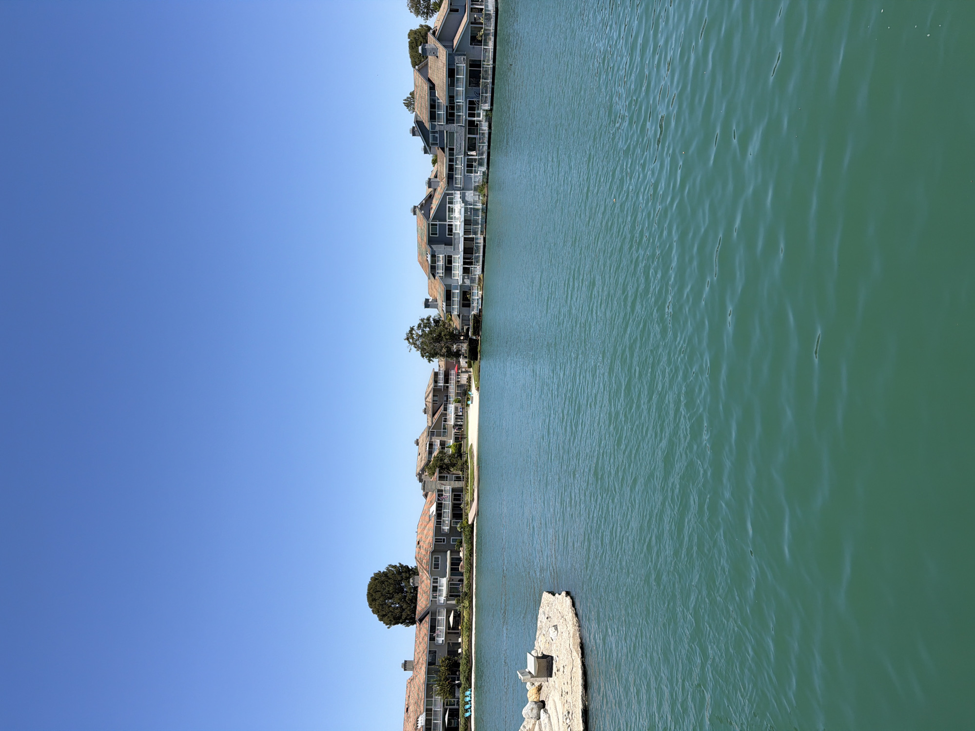 The image depicts a serene waterfront scene in Irvine, California, United States of America. In the foreground, a body of turquoise water stretches across the frame, with gentle ripples suggesting a calm atmosphere. To the left, a small, rocky outcrop juts into the water, featuring a light-colored bench.
Dominating the midground and background is a row of multi-story residential buildings, characterized by their light gray facades, white railings, and terracotta-colored roofs. These buildings are situated along a sandy shoreline, with lush green trees and landscaping interspersed between them. The architecture suggests a modern, planned community.
The sky above is a clear, vibrant blue, indicating a sunny day with excellent visibility. The overall impression is one of tranquility and upscale living, common in coastal or lakeside communities within Irvine. There are no visible people or animals in the frame, emphasizing the peaceful solitude of the setting.