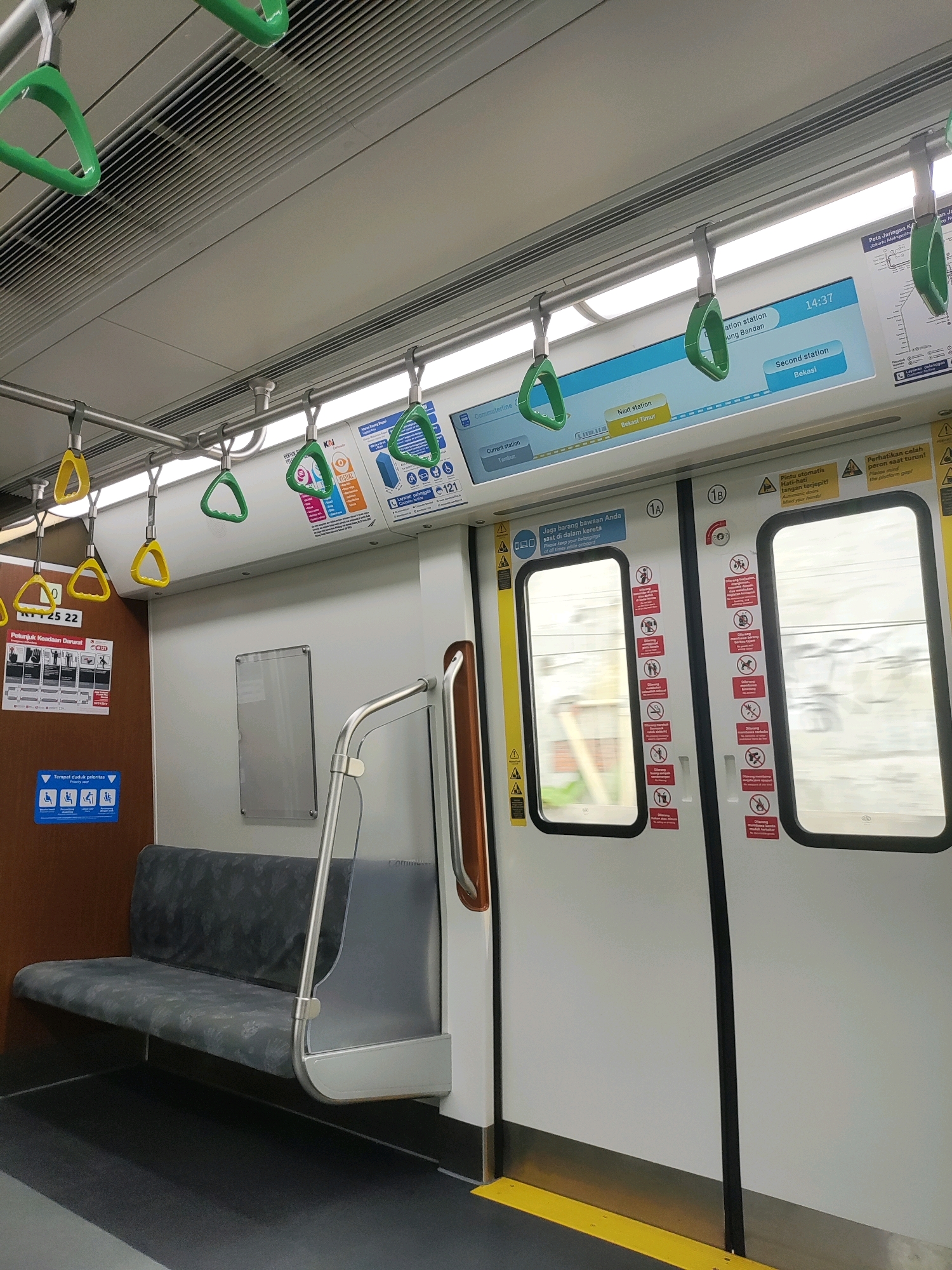 The image depicts the interior of a modern commuter train car. The scene is clean and well-lit, with white walls and grey flooring. Several green and yellow plastic handholds are visible, suspended from a metal rail along the ceiling. These are designed for passengers to hold onto while the train is in motion.
On the left side of the image, there is a grey upholstered bench seat. Next to the seat, on the wall, is a brown panel with several informational signs. One sign, labeled "RT 25 22", is a safety instruction guide with pictograms. Below it, a blue sign indicates "Tempat duduk prioritas" (priority seating) with icons representing pregnant women, elderly individuals, people with disabilities, and parents with young children.
To the right, there are two white train doors, each with a large window. The windows show a blurred view of the outside, indicating the train is moving. Both doors have multiple red and white warning stickers with pictograms, illustrating prohibited actions such as smoking, eating, drinking, and bringing certain items. One door is marked "1A" and the other "1B". Above the doors, a digital screen displays train information, including the current time "14:37", the current station as "Tambun", and the next station as "Bekasi Timur". A second station is listed as "Bekasi".
The overall environment is characteristic of public transportation within the city of Bekasi, Indonesia, given the station names mentioned on the digital display and the Indonesian language on the signs. The scene is empty of passengers, suggesting the photograph was taken at a time when the train is not in heavy use, or between stops. The lighting suggests the interior lights are on, and there is also light coming from the windows.