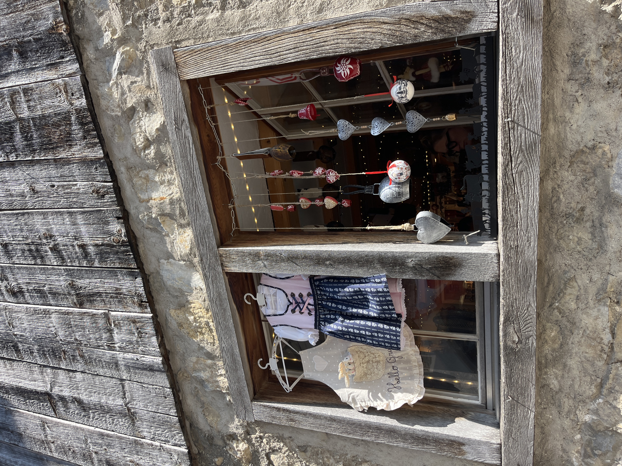 The image shows a shop window with traditional clothing and decorations. On the left, an apron with a heart motif and the words "Hello Gruu" is displayed. Next to it is a pink and blue dirndl, a traditional dress. The window is adorned with various hanging ornaments, including bells, hearts, and round baubles, some featuring Christmas scenes. A silhouette of cows and figures runs along the bottom of the window. The shop appears to be a souvenir or gift shop, likely catering to tourists. The architecture features rough-hewn wood and stone, suggesting a rustic or historic setting. The scene is illuminated by natural sunlight. No specific location in Epagny, Switzerland is identifiable from the image alone.