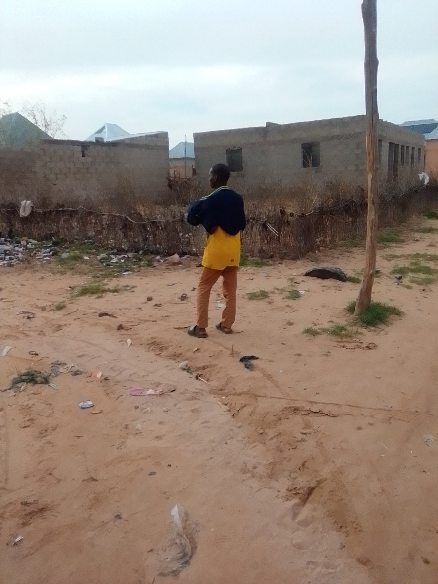 The image depicts a young boy standing in a sandy area in Gombe, Nigeria. He is wearing a navy blue jacket over a yellow t-shirt and brown trousers. Behind him, a wooden pole stands vertically. In the background, there are unfinished concrete buildings with visible brickwork and windows. The ground is covered in sand and scattered debris, including plastic bags, discarded items, and vegetation. The sky is overcast, suggesting a cloudy or hazy day. The overall scene portrays an environment that appears to be an undeveloped or in-progress urban area.