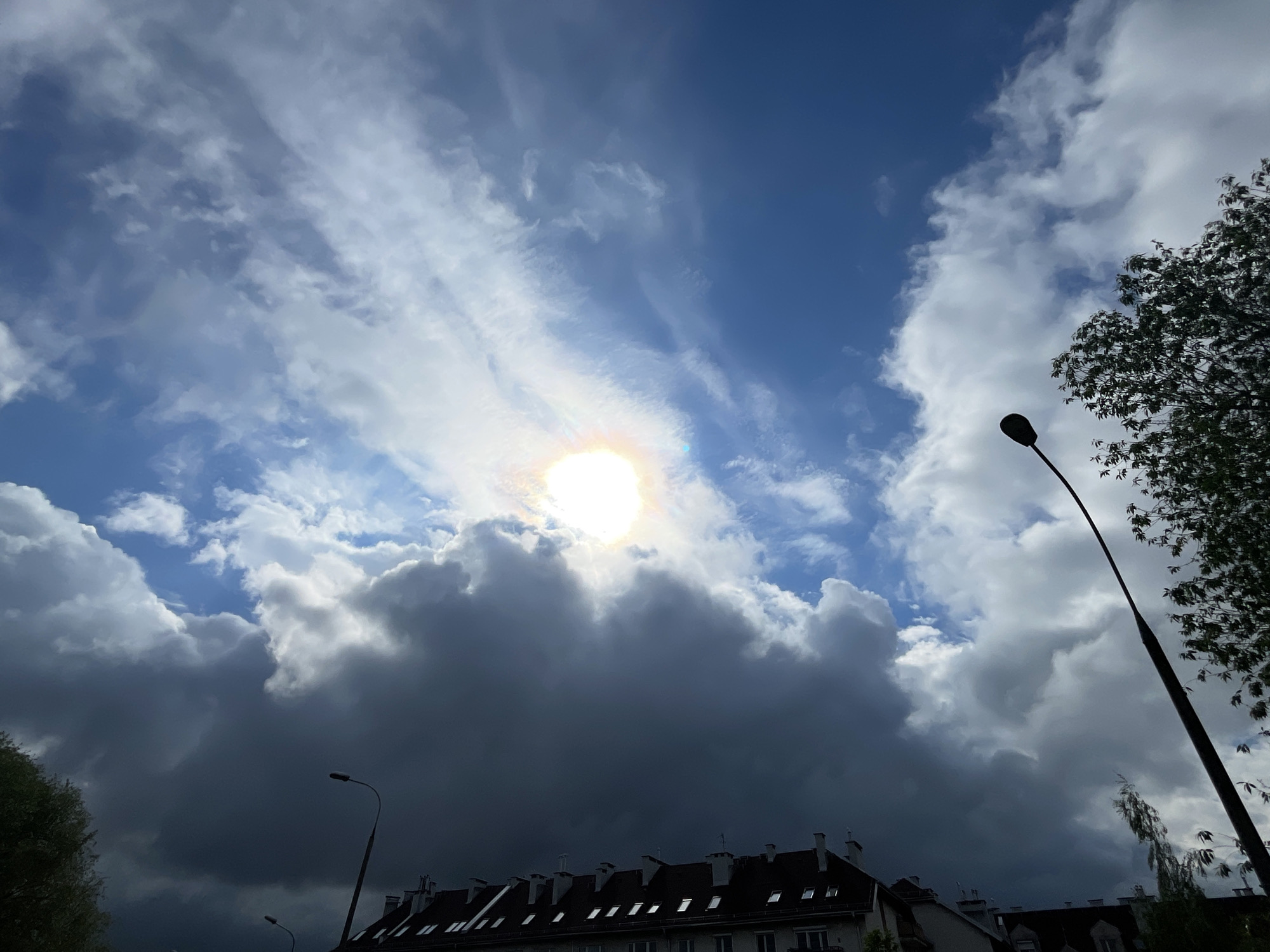 The image depicts a daytime scene with a cloudy sky partially obscuring the sun. The buildings below appear to be residential, with a dark red/brown tiled roof and chimneys visible. Streetlights are present on the lower sides of the shot, and greenery is visible on both sides. The location is not specified in the prompt, so there is no information available to give an exact location or address.