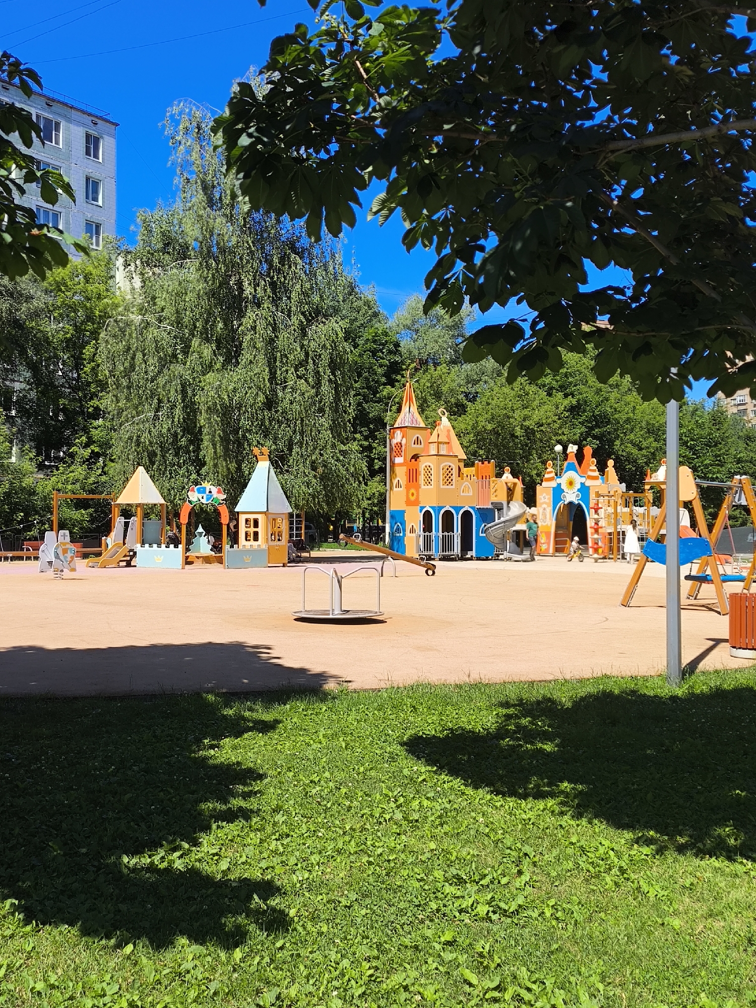 An outdoor shot captures a brightly colored playground on a sunny day. The playground features a large, castle-themed structure with slides, tunnels, and climbing elements in shades of orange, yellow, and blue. To the left, a smaller play area includes a yellow triangular roof structure with slides and what appear to be spring riders. In the center, a small, silver merry-go-round is visible. In the background, lush green trees and a tall, light-colored apartment building are present, suggesting an urban or suburban residential setting. A few children are present on the playground, one riding a small yellow toy vehicle near the castle structure, and another child in white near a swing set. The foreground shows a patch of vibrant green grass with shadows from overhead trees.