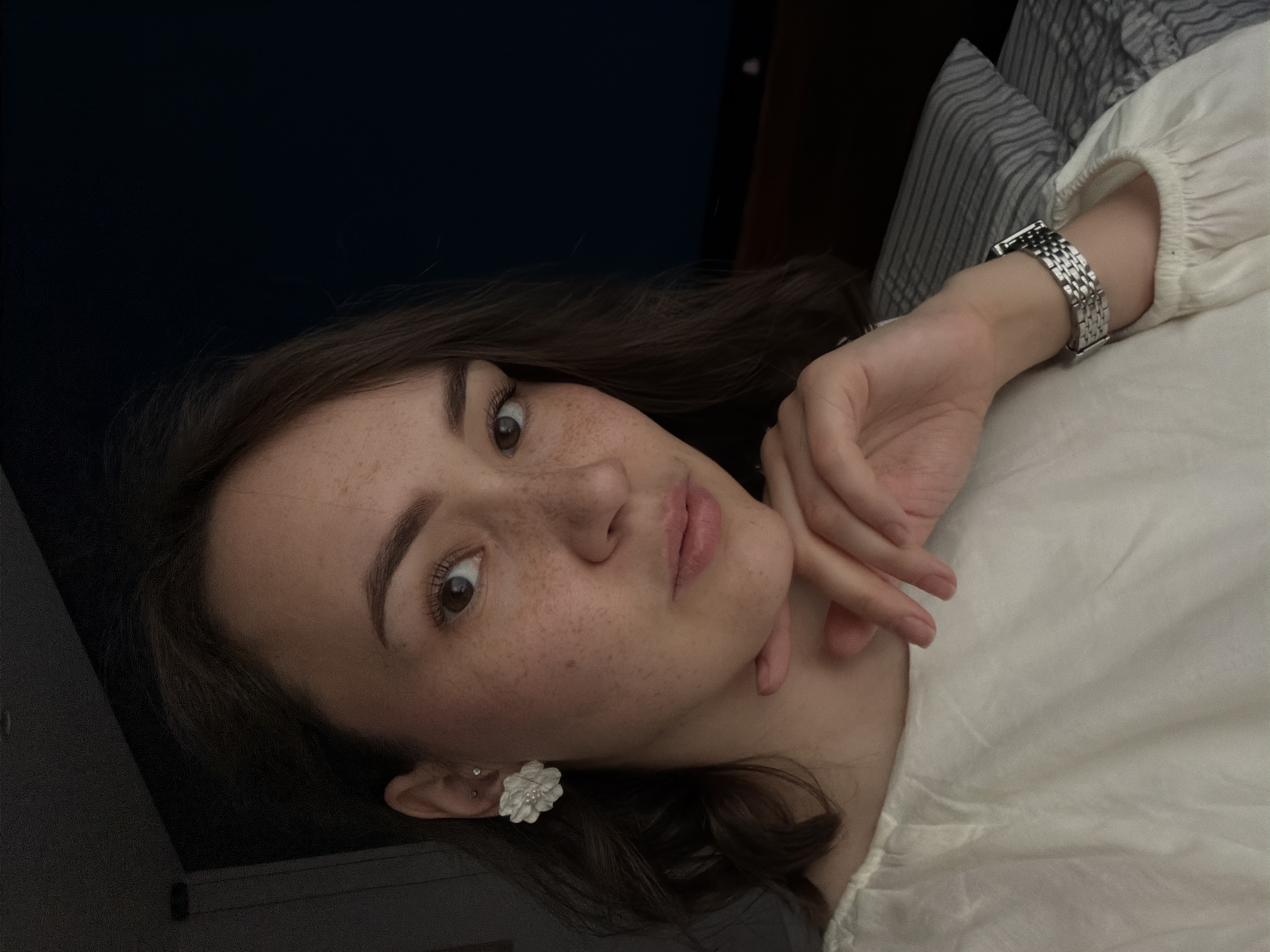 This is a portrait-oriented, low-angle, eye-level shot of a young woman lying on her side, looking towards the camera. Her head is tilted, and her right arm is propped up, with her hand resting near her chin. She has light brown hair that falls around her face and shoulders. Her facial features include large brown eyes, a visible brow line, and full lips. She has a scattering of light brown freckles across her nose and cheeks.
She is wearing a white, long-sleeved top with gathered fabric at the cuffs. A silver, multi-link watch with a rectangular face is visible on her left wrist. She is also wearing a white flower-shaped earring in her left ear, with a smaller stud earring above it.
The background is dimly lit. To the left, there is a dark, possibly upholstered surface. To the right, there is a pillow with thin gray and white stripes, and a plain, light-colored surface, likely a bedsheet. The overall lighting suggests a low-light environment, possibly indoors in the evening or on a cloudy day. There is no visible text in the image.