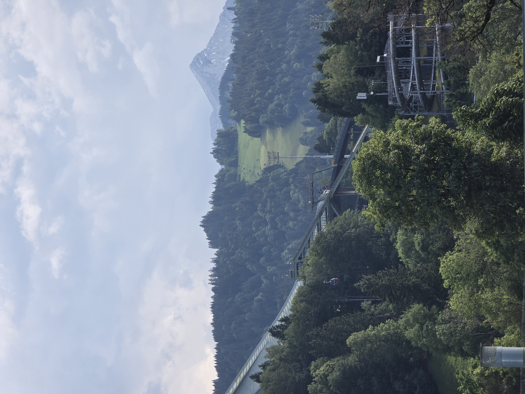 This is a vertical, eye-level, medium shot of a mountainous landscape with a bridge and forest. The sky is partly cloudy and a light blue. In the distance, a snow-capped mountain rises above a densely forested hill. A bright green field is visible on the side of the hill. A large, modern suspension bridge spans across the frame, with its concrete supports and steel cables clearly visible. The bridge is covered in green foliage, and a train can be seen traveling across it. To the right of the bridge, a forest of green trees covers the landscape. A cable car station with several gondolas is visible within the trees on the right side of the frame. The scene is bathed in natural light, suggesting it is daytime. The location cues and the style of the bridge and cable car suggest a European Alpine region, consistent with Garmisch-Partenkirchen, Germany.