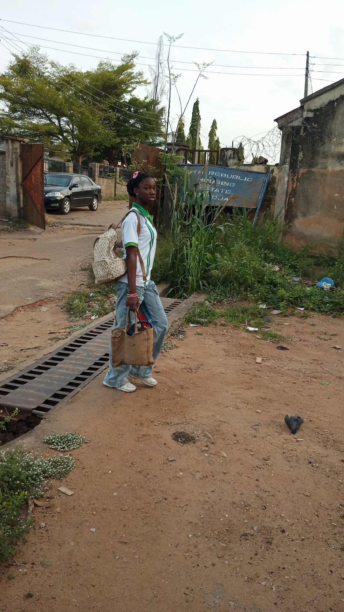 A young woman stands on a dirt road in Adankolo, Nigeria. She wears a white t-shirt with green trim, light-wash jeans, and white sneakers. A backpack is slung over her left shoulder, and she carries a tan tote bag in her left hand. To her left is a storm drain, and a black car is parked beyond an open gate. Behind her is a concrete wall topped with barbed wire. A sign partially obscured by vegetation reads "South Republic Housing Estate, Oja." Trash is scattered on the ground.