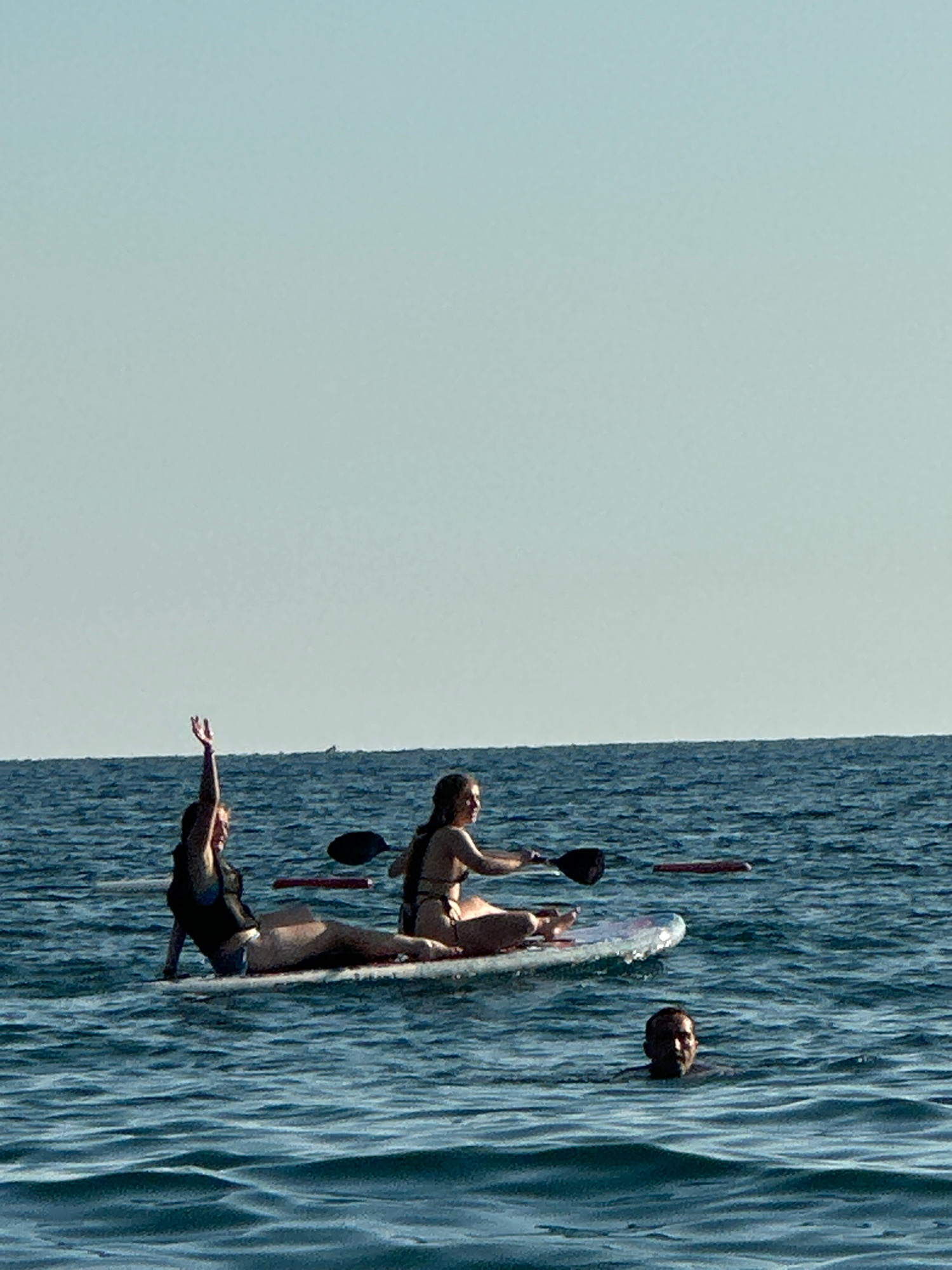 In the open water near Boreti, Montenegro, two individuals are engaged in paddleboarding on a clear, sunny day. The sea is a deep blue, with gentle waves creating a textured surface. The sky above is a pale, cloudless blue.
On a paddleboard, one person is positioned towards the front, sitting with their legs extended. They are wearing a dark-colored top and lighter-colored bottoms. Their right arm is raised in the air, with their hand open.
Behind them on the same paddleboard, another individual is seated, facing towards the right. They are wearing a bikini. They are holding a paddle, with the paddle blade resting on the water.
In the foreground, a third person's head is visible above the water, looking towards the camera. Their face is dark due to shadow.
The scene suggests a recreational activity taking place in a coastal area.