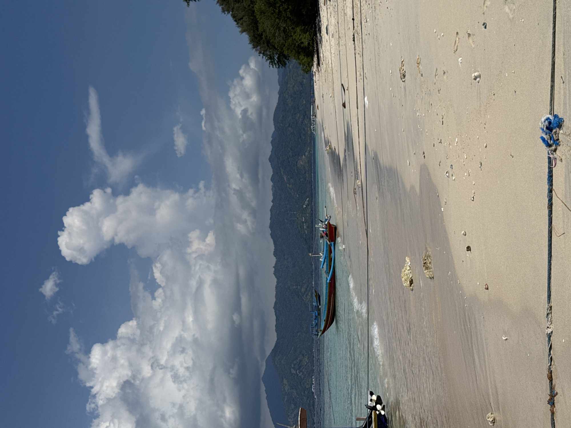 The image shows a beach scene in an unknown city in Indonesia. The sky is predominantly blue with large, white cumulus clouds. A mountain range covered in lush green vegetation forms a backdrop. The beach is sandy, with several traditional Indonesian fishing boats resting on the shore, partially submerged in the clear turquoise water. The sand is marked with patterns of waves and scattered debris. A rope with a blue knot is visible in the foreground.
