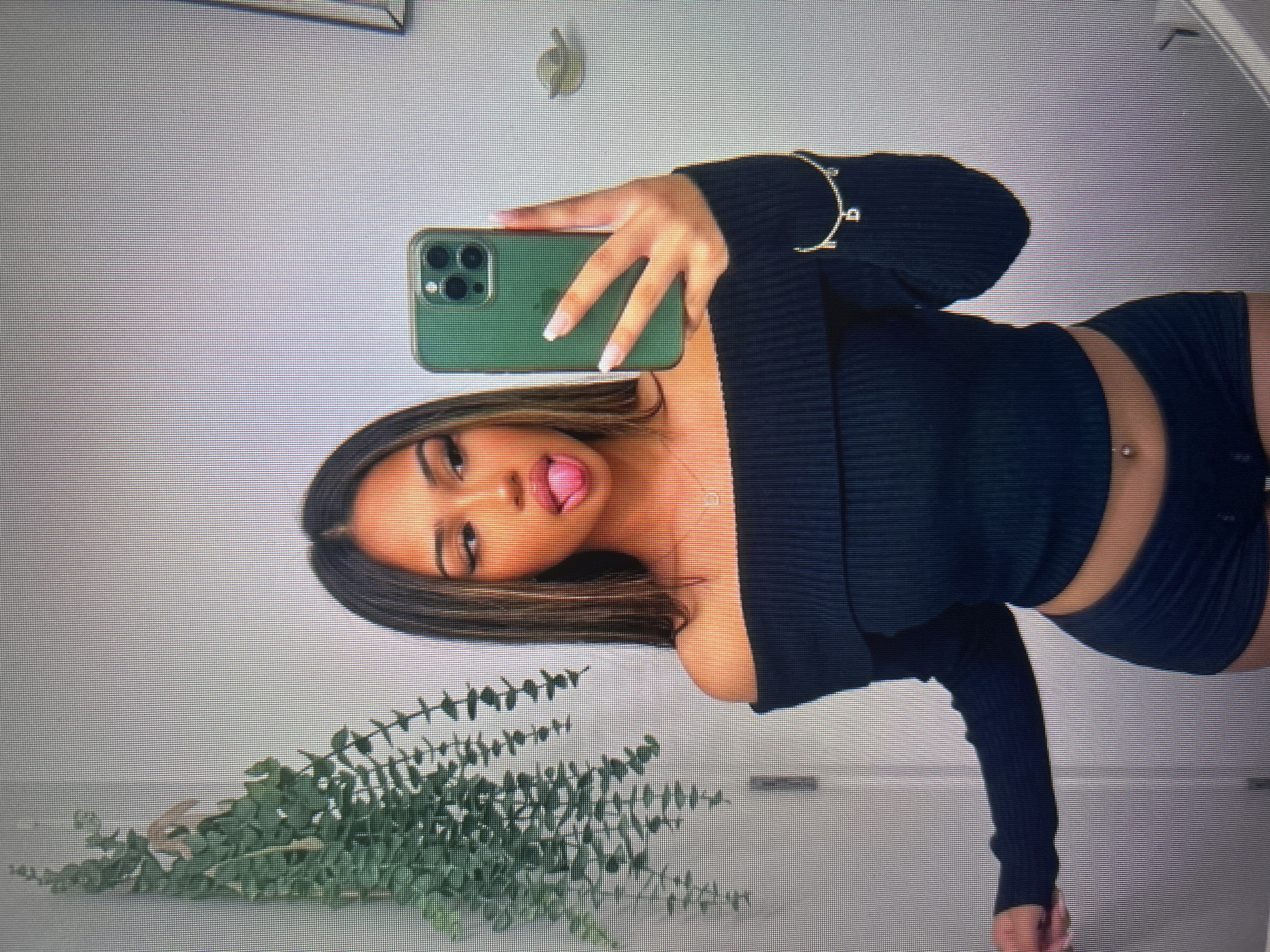 A young woman with dark, highlighted hair is taking a mirror selfie in what appears to be a bathroom. She is wearing a black off-the-shoulder top and dark shorts. She has a green phone in her right hand, taking the picture. Her tongue is sticking out playfully. A green plant hangs to the left of the frame. There is a delicate bracelet with a letter charm on her left wrist. The environment is simple, with a white tiled wall visible behind her. There are no specific identifying features that place the image in Stara Zagora, Bulgaria.