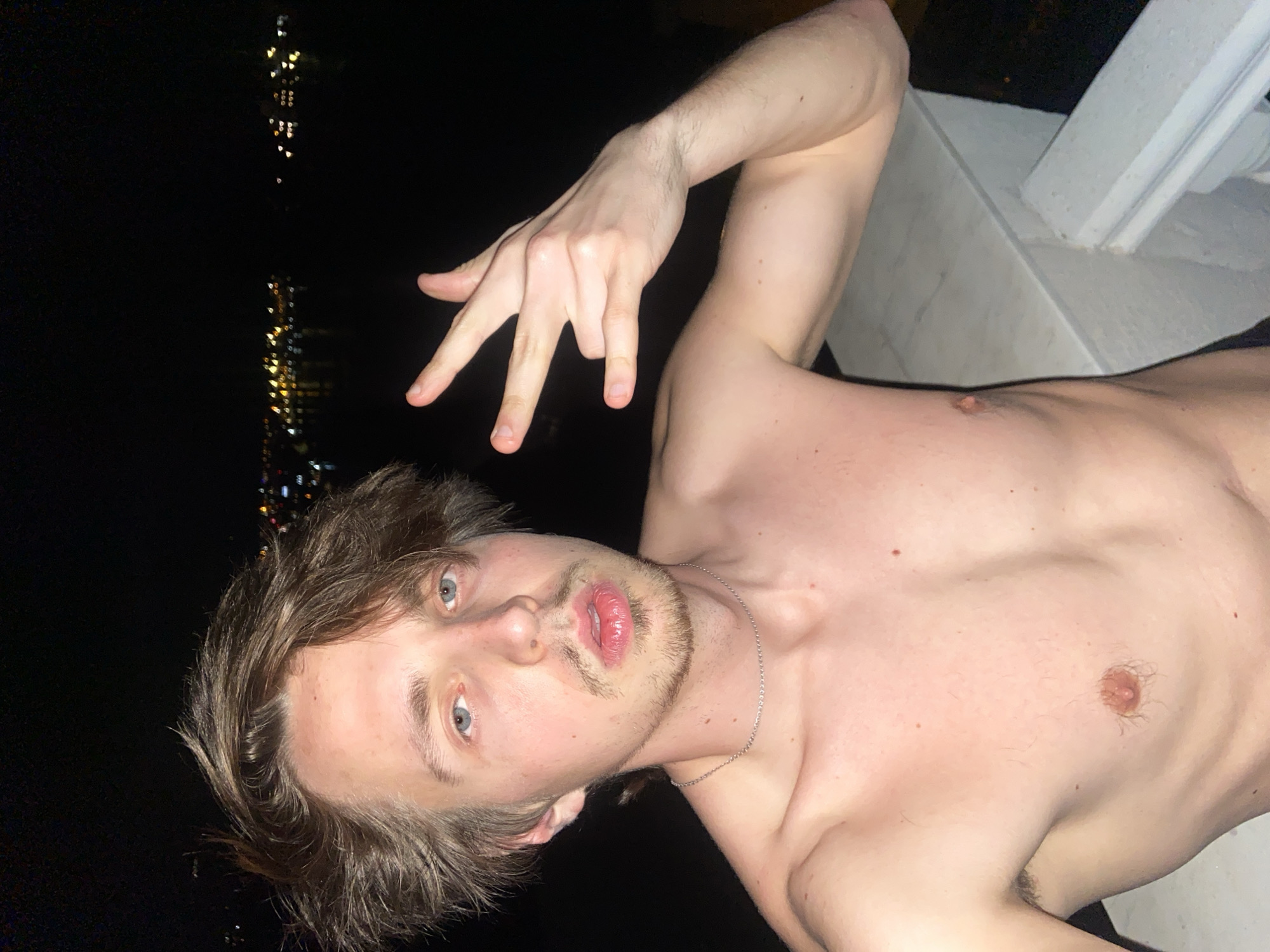 A shirtless Caucasian male is posing outdoors at night in Šušanj, Montenegro. He has light brown hair and blue eyes. He is making a hand gesture with his left hand. A silver chain is visible around his neck. Blurry lights are visible in the background, possibly from buildings in the distance. A white column is partially visible on the right side of the image.