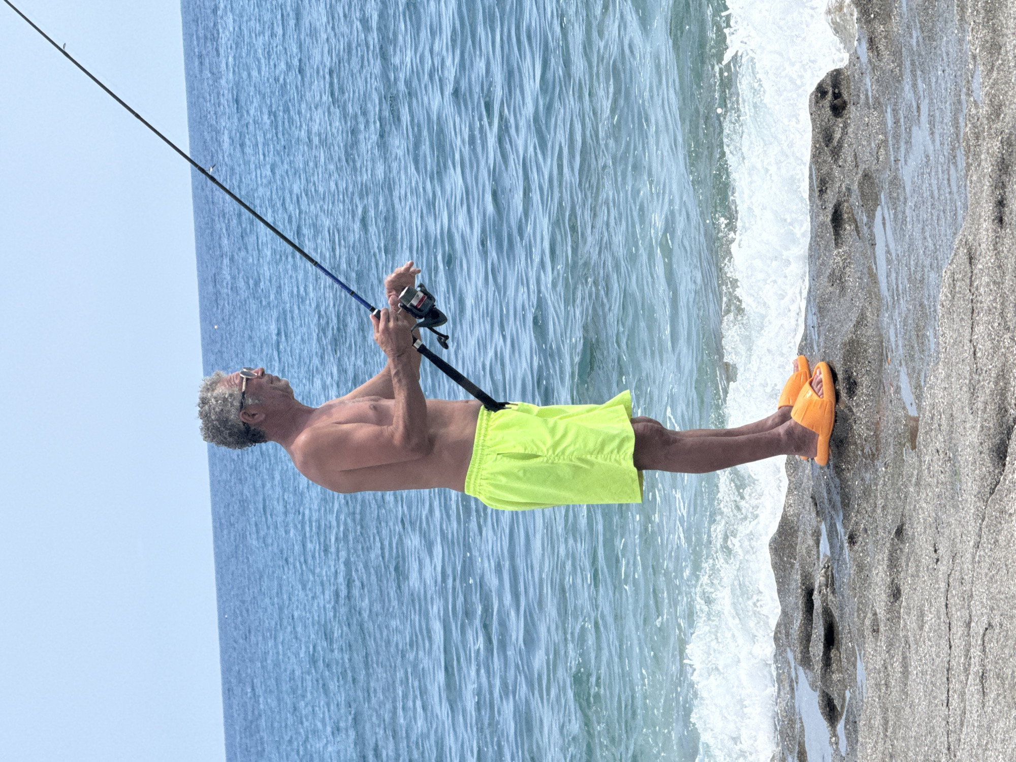 A shirtless man with curly gray hair and sunglasses is fishing on the rocky shore of Alanya, Turkey. He is wearing bright yellow swim trunks and orange flip-flops. The man is holding a fishing rod and reel, casting into the blue ocean. Waves crash against the rocks, creating white foam. The sky is clear and blue.