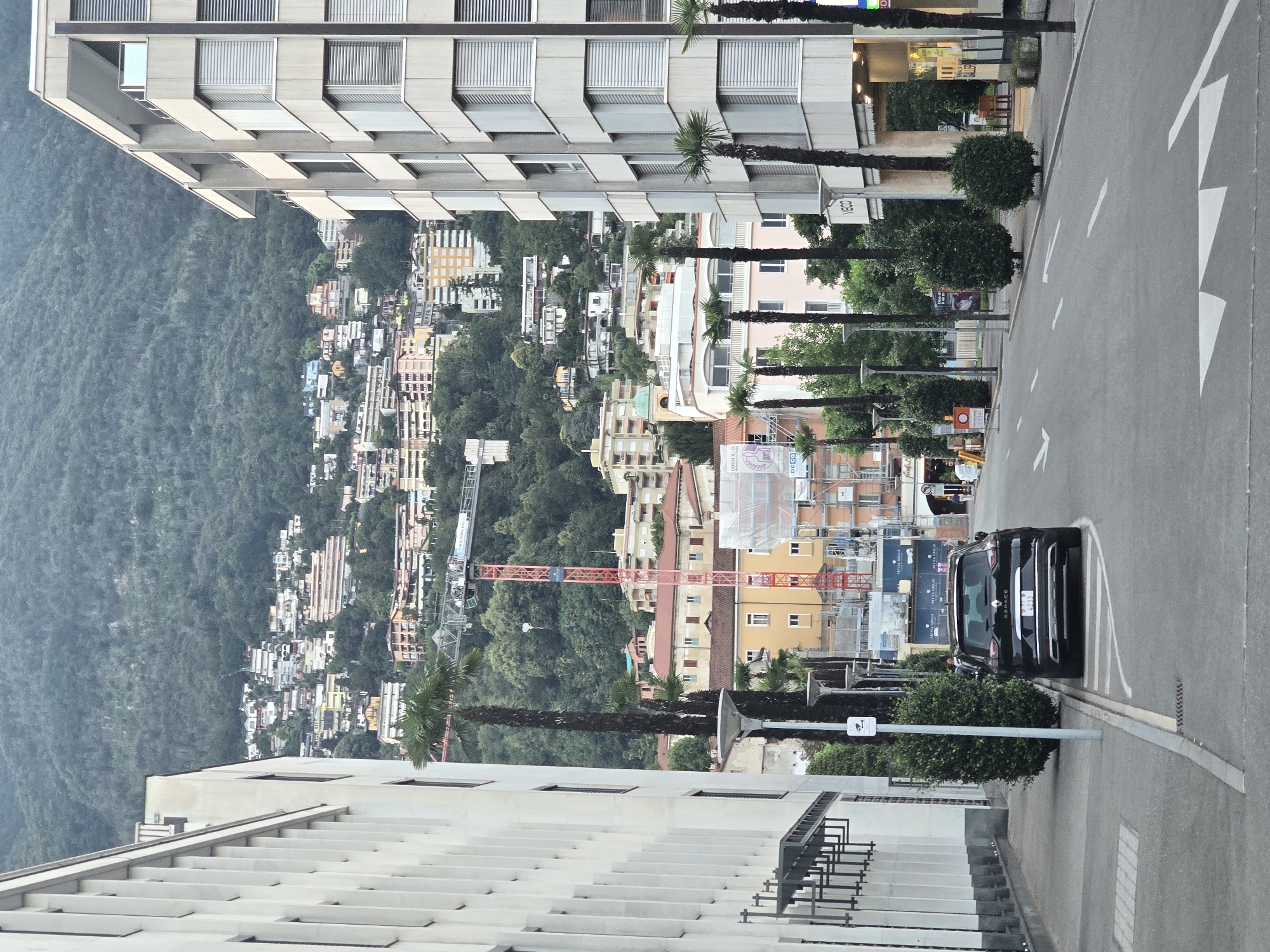 The image depicts a street scene in Locarno, Switzerland, with a modern building on the left and a multi-story building on the right, facing a hill densely covered with residential buildings and trees. A black SUV is parked on the right side of the road. The road has lane markings and a pedestrian crossing. Several palm trees line the sidewalk. In the background, a construction crane is visible, suggesting ongoing development. The sky is overcast.