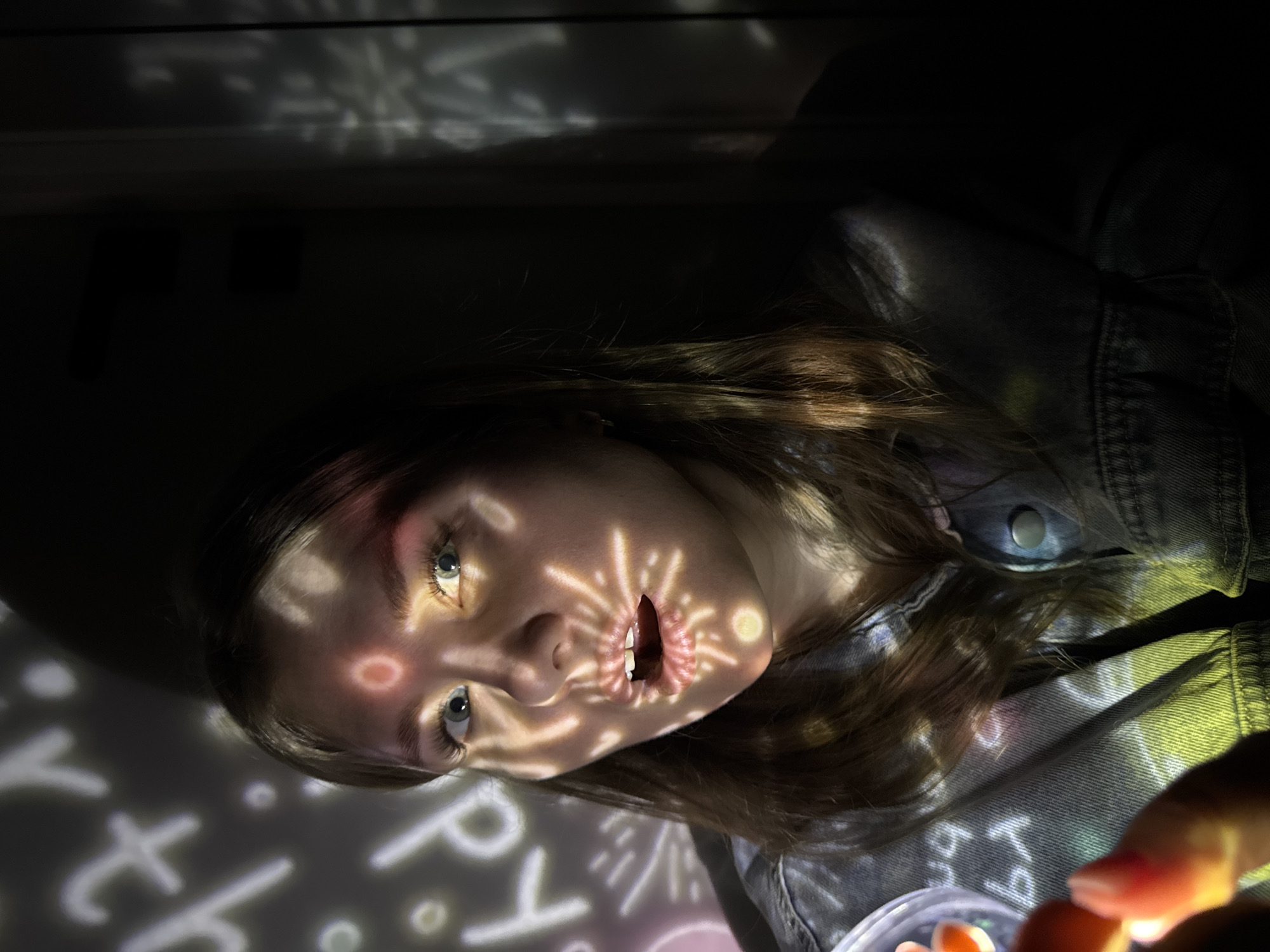This image, located in Gornyi (Dvurechenskoe S/P), Russia, features a young adult female subject within a dark interior space. The subject, possessing light-colored eyes and brown hair, is clad in a denim jacket and looks directly ahead with a slightly open mouth. Her face and the adjacent dark wall are partially covered by projected light patterns. These patterns include white numbers, abstract shapes, and starbursts, rendered in white, yellow, red, and green. Specifically, a red circular pattern is visible on her forehead, and a yellow starburst pattern illuminates her chin. In the lower-left foreground, a hand, consistent with the subject's, holds a translucent object emitting an orange-red luminescence. The background consists of dark, featureless wall surfaces, with a vertical line visible on the right.