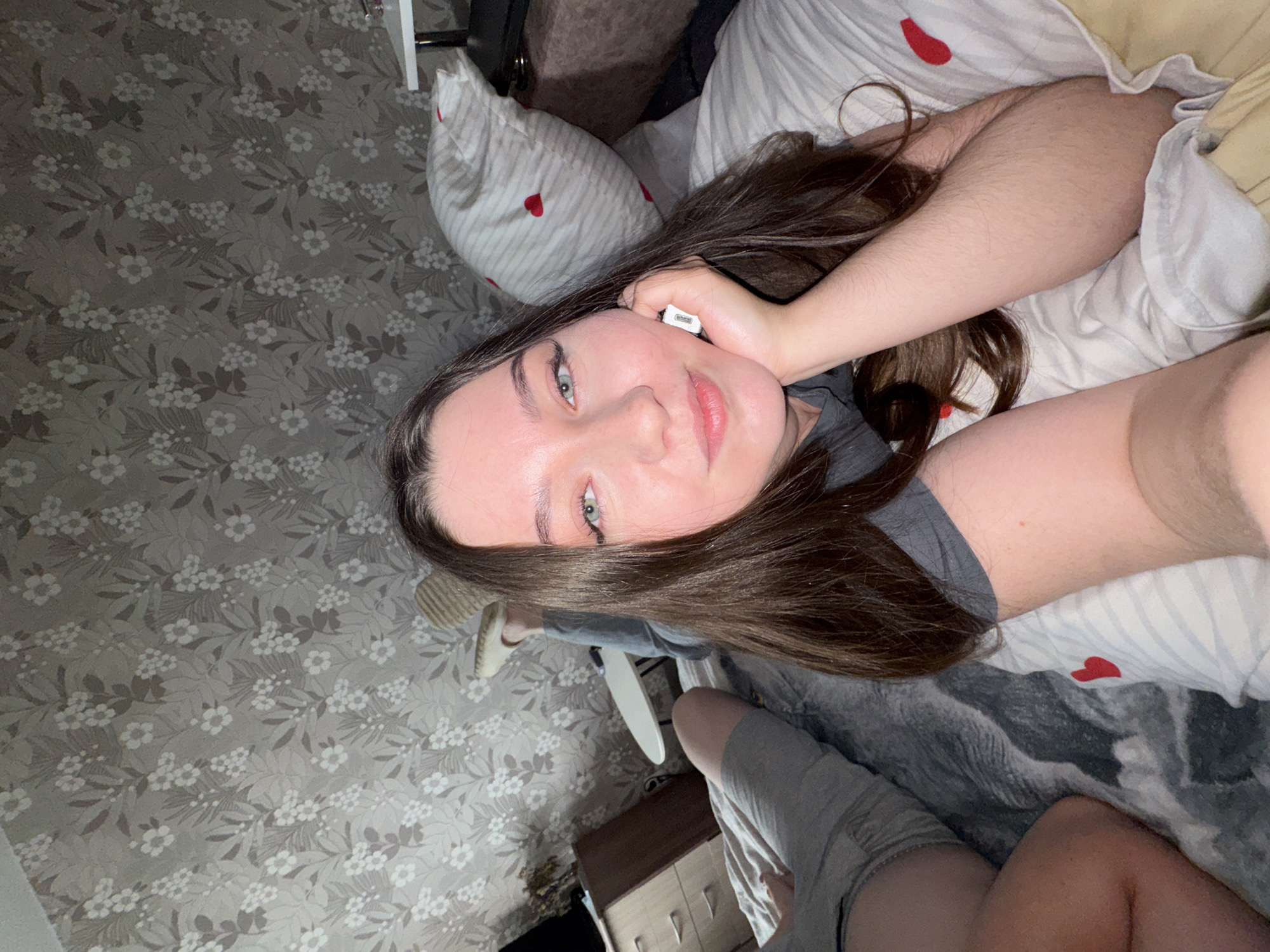 The image shows a young woman lying on a bed in a room with floral wallpaper. She is looking at the camera with a slight smile, resting her head on her hand. She is wearing a grey t-shirt and grey shorts. The bed has a striped duvet with small red hearts. A straw hat is visible on one of her feet. A bedside table with drawers is in the background, along with a television. The room appears to be a bedroom in a residential setting. The location is identified as Bashkultaevo, Russia.