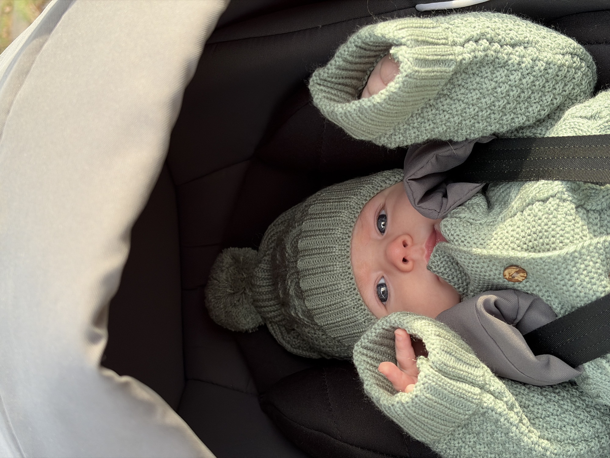 A baby, secured in a black stroller with a harness, is lying on their back. The baby is wearing a green knitted outfit with a matching hat adorned with a pom-pom. The baby's hands are visible, with the left hand positioned near their face and the right hand near their torso. The stroller's interior is dark, contrasting with the stroller's light-colored hood on the left side of the frame. The scene appears to be indoors. The location is not discernible from the image.