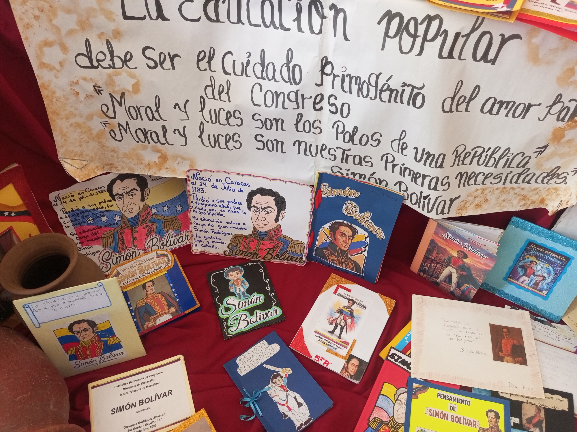 The image depicts a display featuring the life and legacy of Simón Bolívar. A large, handwritten banner with a quote attributed to Bolívar is prominent, stating, "La educación es el cuidado del Congreso. Moral y luces son los polos de una República; primeras necesidades." Below this, various handmade books, posters, and a clay pot are arranged on a red fabric surface. Many of the items are adorned with images of Bolívar and Venezuelan flags, indicating a patriotic theme. One of the visible book titles reads "Simón Bolívar." A document with the header "República Bolivariana de Venezuela. Ministerio de Educación. U.E.B. 'Victoria de Matasiete'. Simón Bolívar (Breve reseña)" suggests a connection to education and a specific school in Santa Ana, Venezuela, likely from a student project. The overall scene appears to be an exhibit or a classroom presentation. There are no people visible in the image.