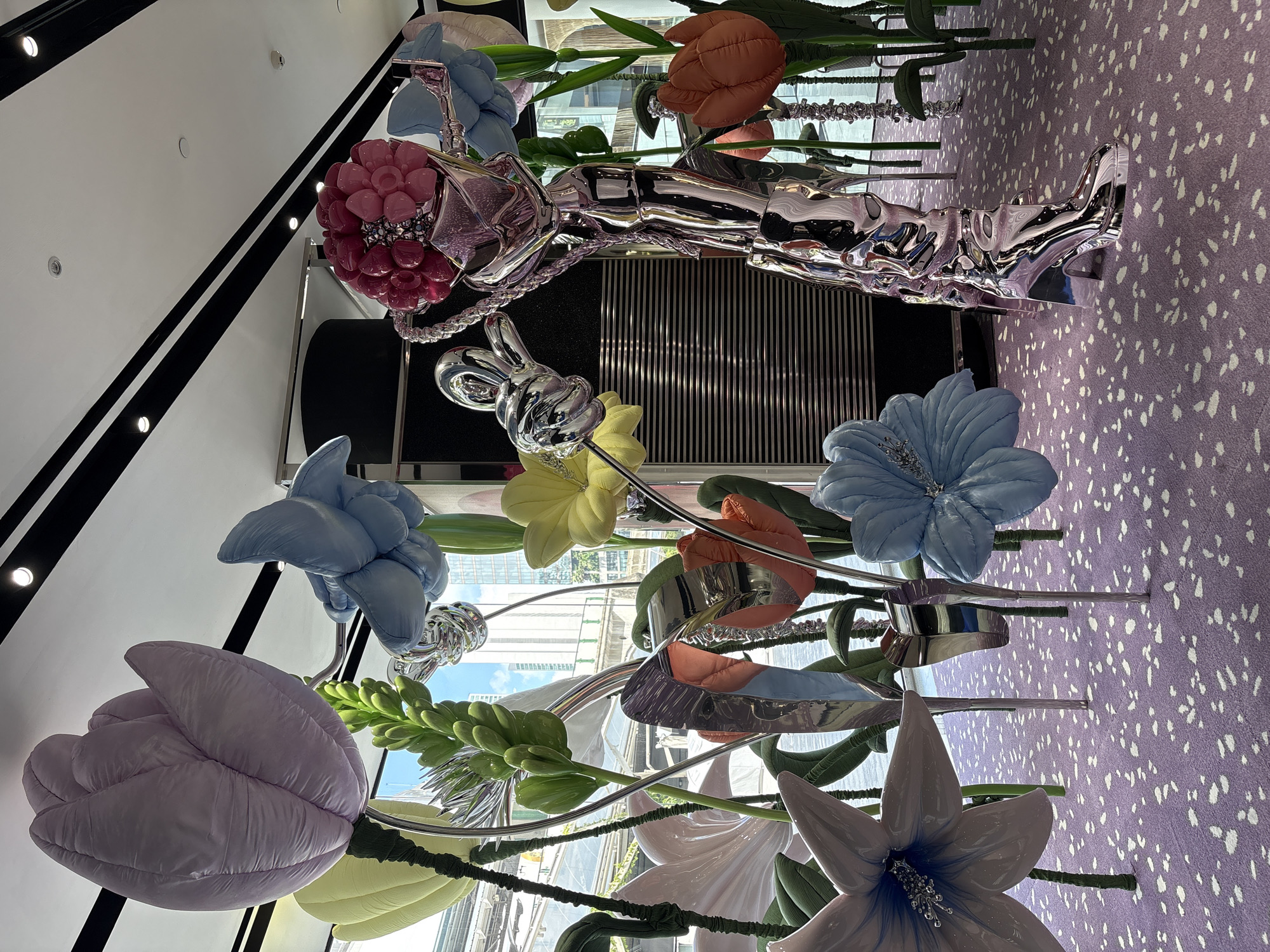 An interior shot of a display at the Siam Paragon shopping mall in Bangkok, Thailand. It features an assortment of oversized, puffy, fabric flowers in pastel colors, including lavender, pale blue, yellow, orange, and white. A polished chrome mannequin is posed horizontally across the middle of the image. The foreground is dominated by the stems of the fabric flowers and the background shows a patterned carpet. Light enters from a window reflected throughout the scene.