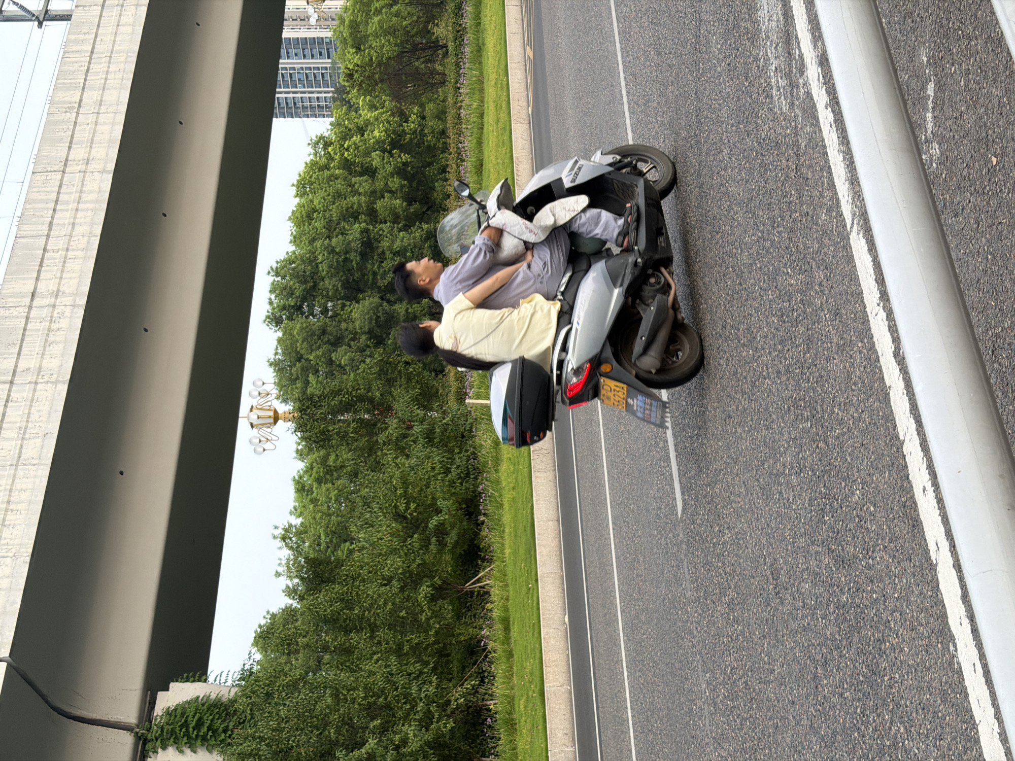 In Jiangjin, China, a man and a woman are riding a silver scooter on a paved road. The man is driving, wearing a grey outfit and black sandals. The woman is riding as a passenger, wearing a pale yellow top and dark pants, with her arm around the man. They have a black storage box mounted on the back of the scooter and a light-colored blanket draped over the front. The scooter has a yellow license plate with the characters "6623A" visible. The road is bordered by a grassy median with some small purple flowers and a row of lush green trees. In the background, a tall building stands to the right, and a large concrete overpass with overhead wires stretches across the top of the frame. The sky is overcast.