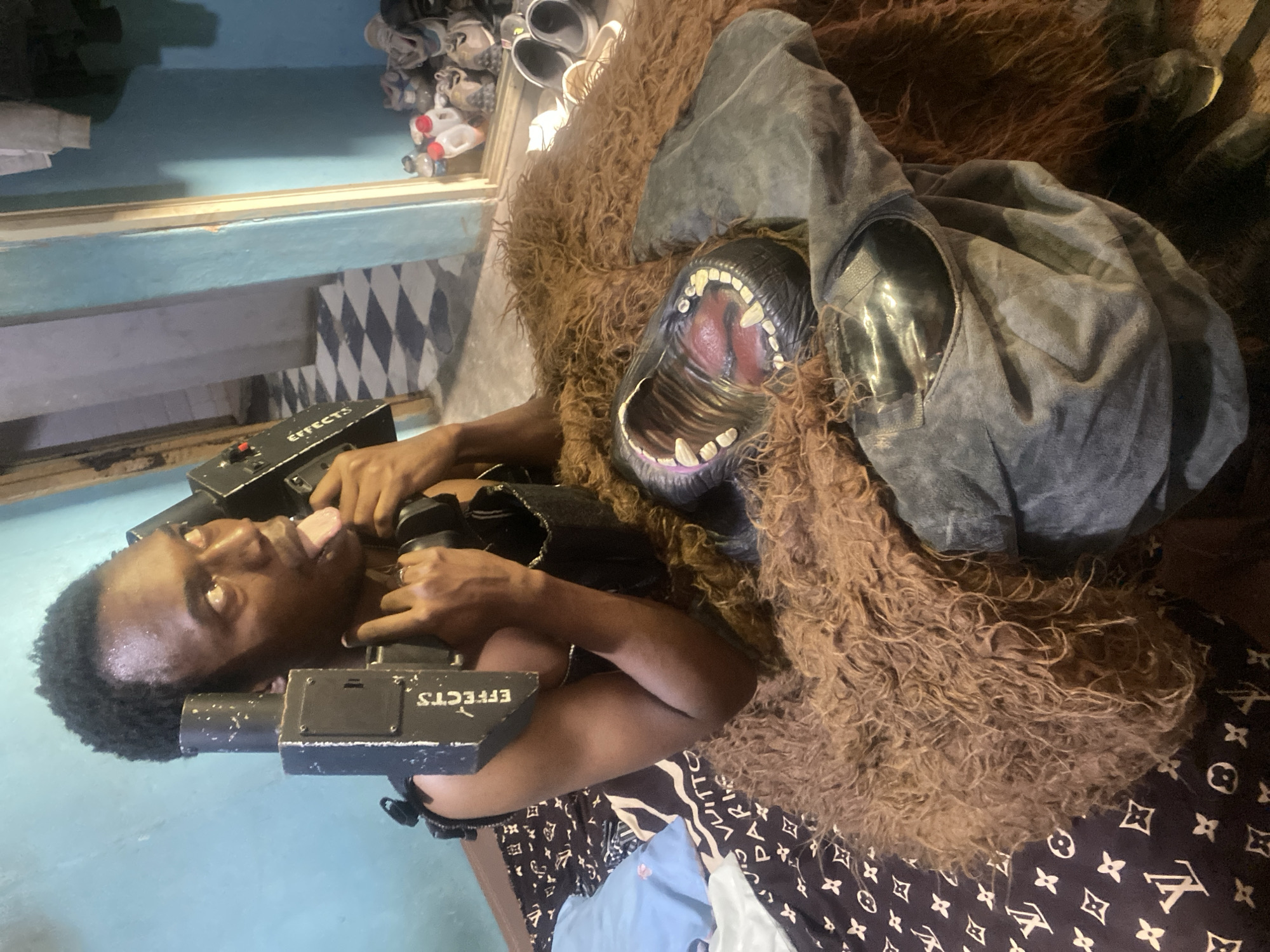 An indoor scene in Jos, Nigeria, features a young Black man posing next to a large, shaggy brown gorilla or beast costume. The man, with short, curly dark hair and wearing a dark top, holds two black prop devices, labeled "EFFECTS," one in each hand. He has an exaggerated expression, with his eyes rolled back and tongue sticking out, suggesting a playful or humorous intent.
The costume head, positioned to his right, has an open mouth revealing prominent white teeth and a red-pink interior. Long, wavy brown fur covers the costume, with a grey fabric garment draped over part of it. The man and costume are resting on a fabric with a repeating pattern resembling the Louis Vuitton monogram.
In the background, a blue wall with a lighter blue horizontal strip is visible. A patterned floor with black and white diamonds can be seen on the left. On a shelf or elevated surface in the upper background, there are several pairs of shoes and multiple plastic bottles. The setting appears to be an informal space, possibly a room used for creative projects or staging.