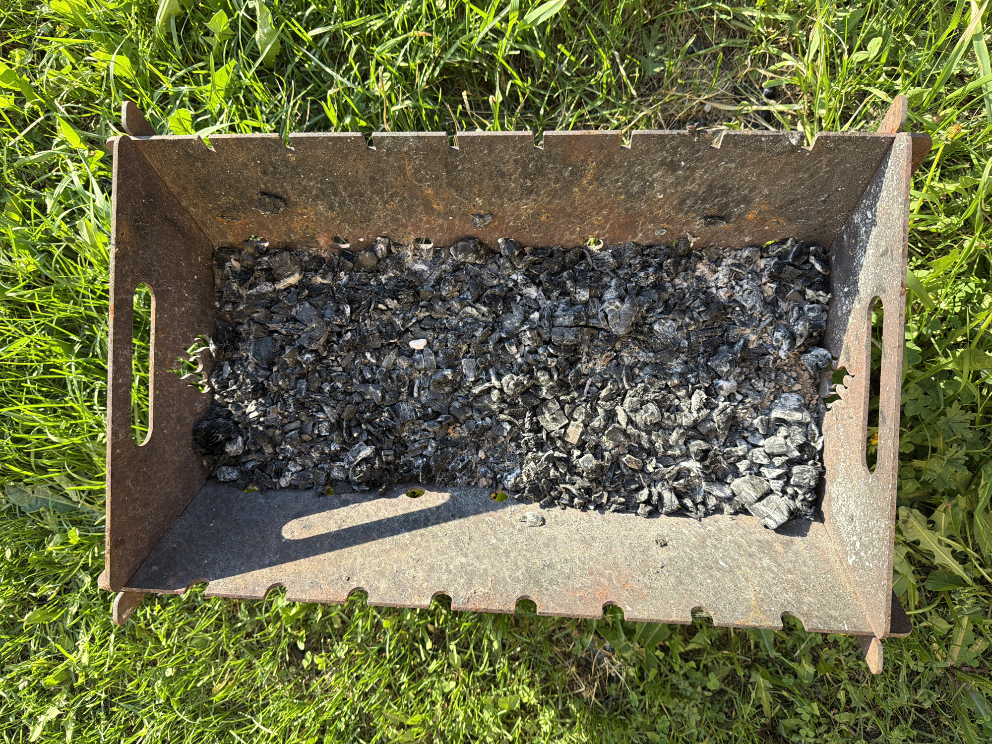 The image depicts a rectangular, rusty, metal grill filled with charcoal remnants. The grill is positioned on a lawn of green grass, likely outdoors. Bolsheustyikinskoye, Russia is not discernible from the image.