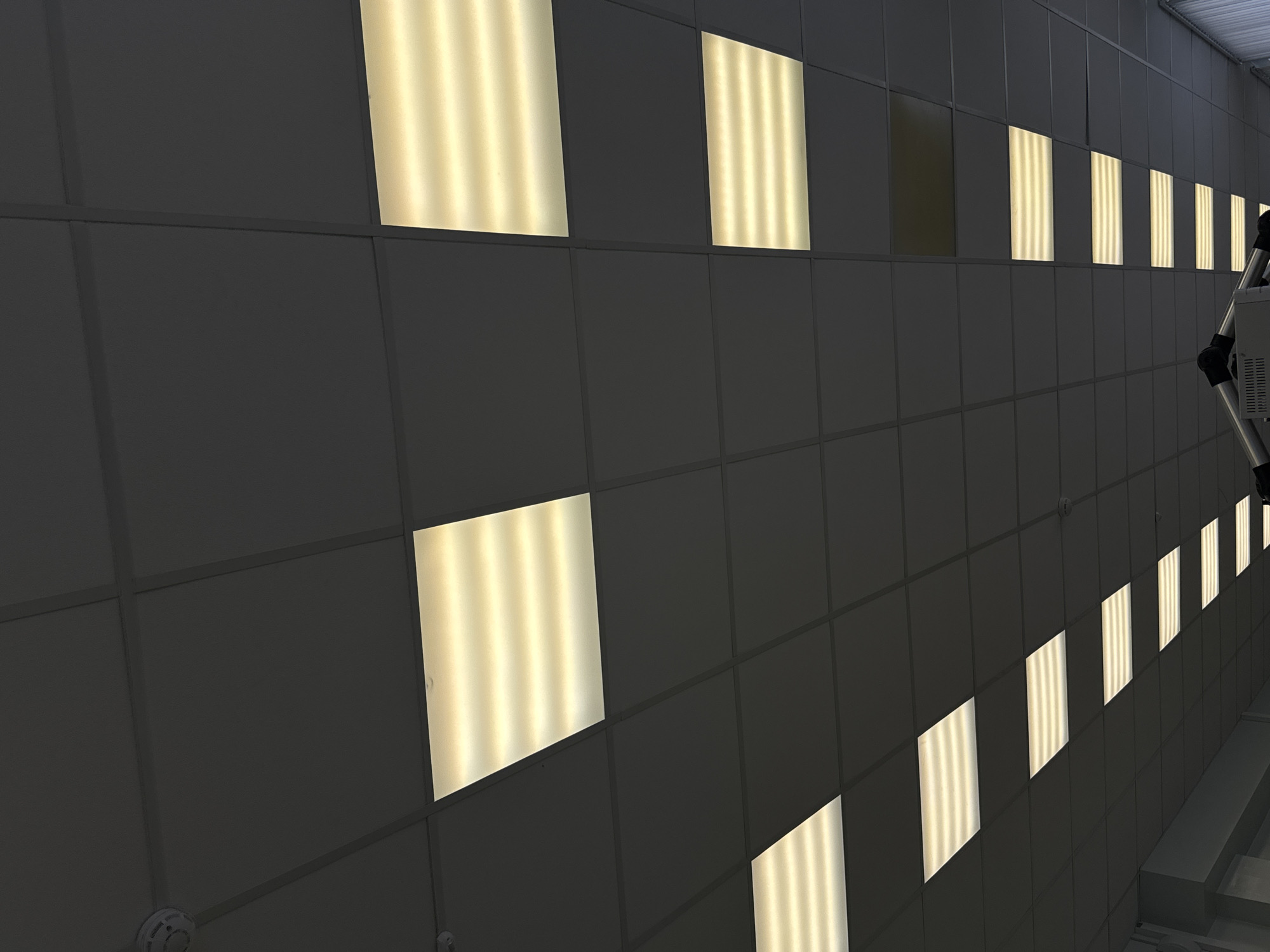 The image shows a ceiling of gray square panels with recessed fluorescent lights, some illuminated and some unlit. The lights create a geometric pattern. A ladder is partially visible on the right side of the image. The location is unspecified, and there is no indication of Perm, Russia.