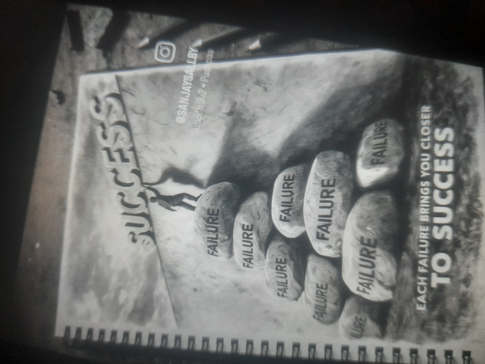 The image is a black and white photograph of a notebook or journal. The cover features the word "SUCCESS" in large letters. Below, a graphic depicts a person climbing stones labeled "FAILURE". The text "EACH FAILURE BRINGS YOU CLOSER TO SUCCESS" is printed nearby. The upper part of the cover also includes the text "@SANJAYSALLBY, Subodh Su2- Pushpaa" and an Instagram icon. The notebook has a spiral binding. The notebook is on a surface. No location is specified.