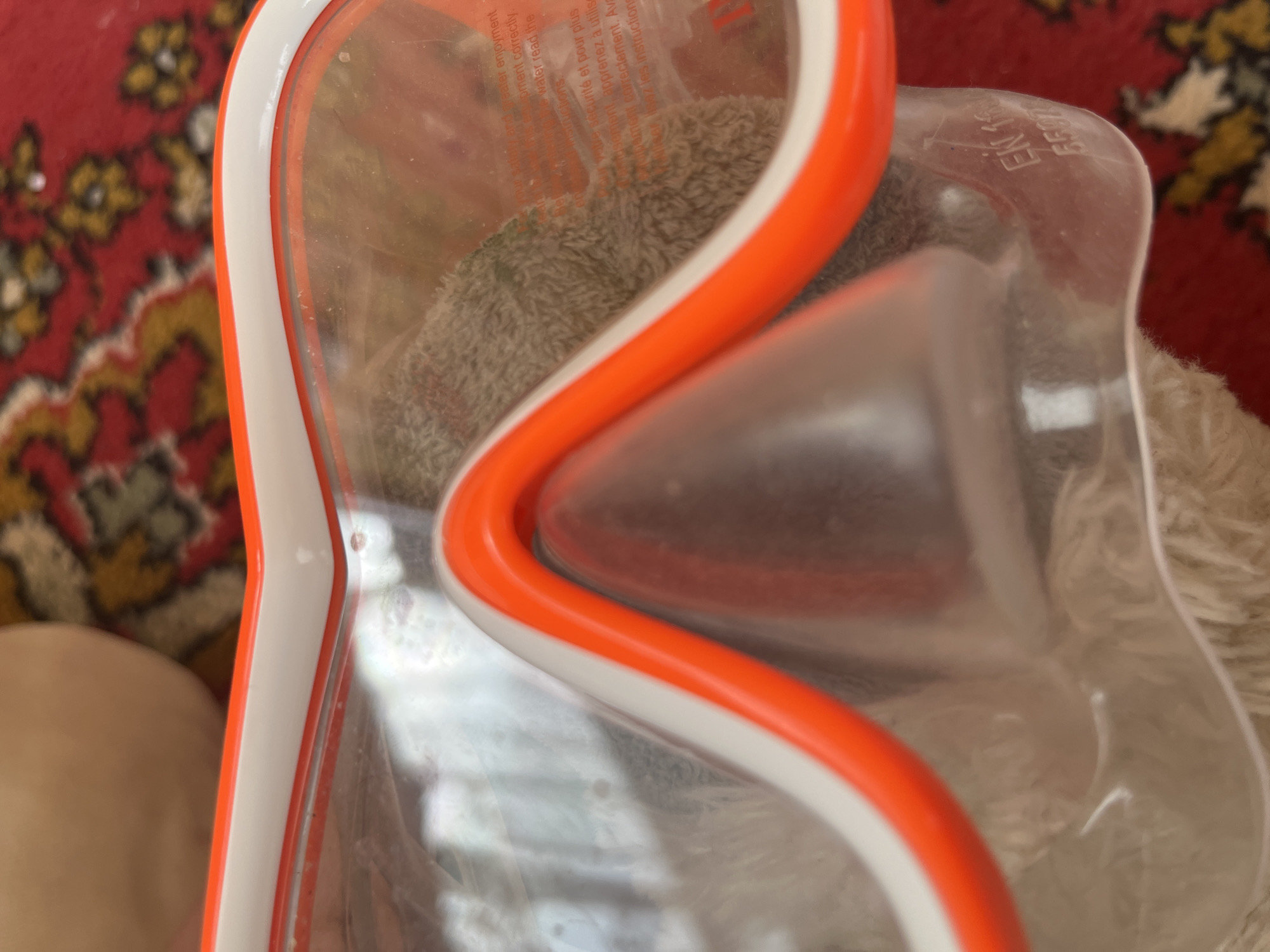 A close-up, slightly elevated perspective depicts the central portion of a swimming or diving mask. The mask features a transparent, slightly smudged plastic lens framed by a rigid white outer rim and a flexible orange inner seal. The nose pocket, also transparent, is visible at the bottom center.
Through the clear lens, text is visible, indicating warning or instructional messages in both English and French. Portions of the English text include "water enjoyment" and "read the instructions," while the French text shows phrases such as "sécurité et pour plus" and "lisez les instructions." Beneath the mask and partially visible through the lens, a soft, light brown or tan terry cloth-like fabric is present, possibly a towel or blanket.
The mask rests on a densely patterned textile surface, likely a rug or carpet, predominantly red with repeating floral or geometric motifs in yellow, gold, and dark tones. On the lower right side of the clear nose pocket, faint embossed characters "EN 16" and "5507" or similar digits are discernible, likely indicating a product standard and/or model number.
The scene does not contain any visual identifiers pertaining to a specific urban environment or country, including the city of Tyumen, Russia.