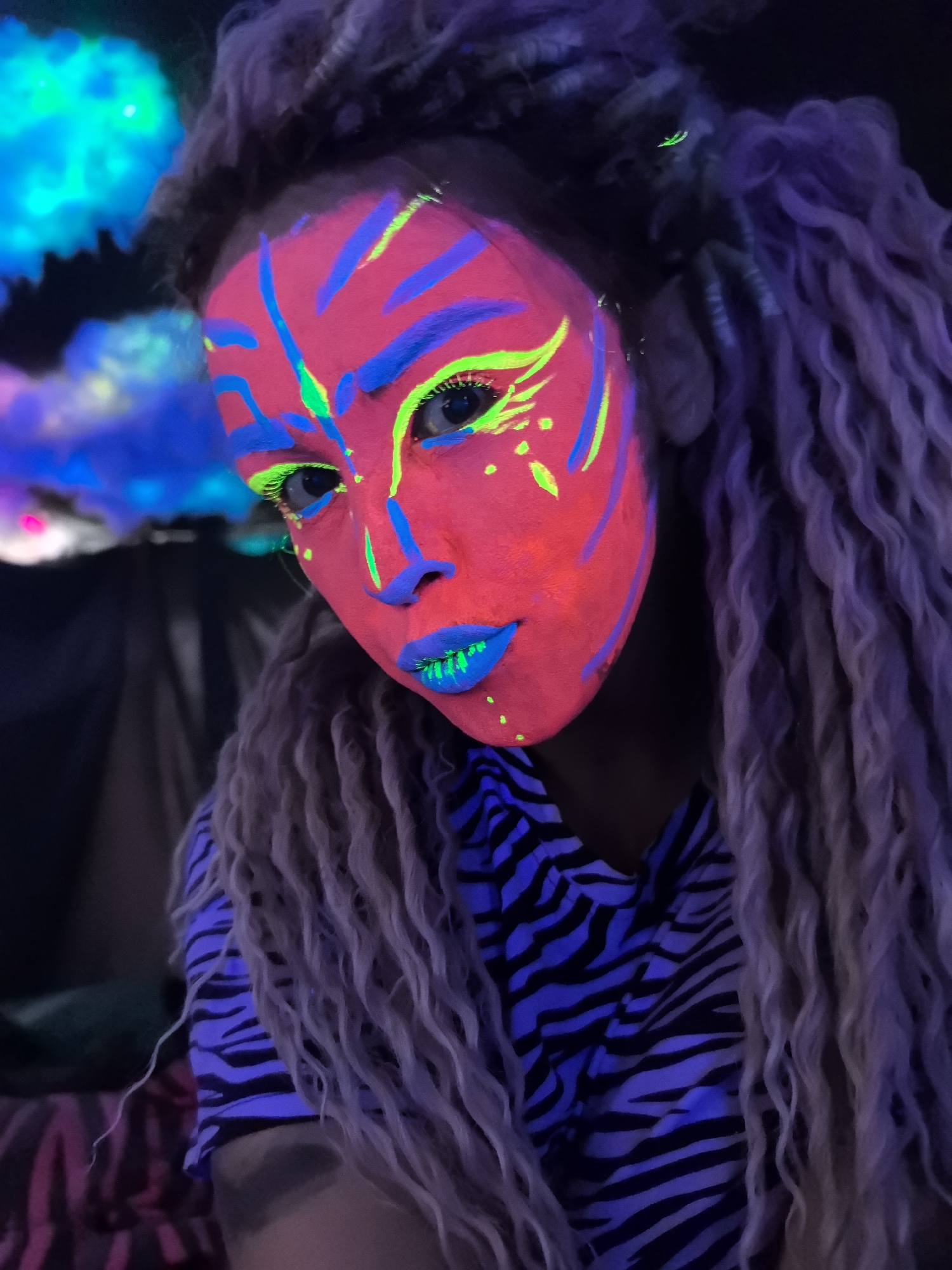 A close-up shot depicts a person with neon makeup in a dimly lit environment. The individual's face is predominantly red, with blue stripes on the forehead and cheeks, green accents around the eyes, and bright blue lipstick. The person has long, curly hair that appears to be a light brown or blonde color. They wear a black and blue zebra-striped shirt. The background shows indistinct, illuminated shapes that suggest an artificial or artistic setting. The location is not discernible within the scene.