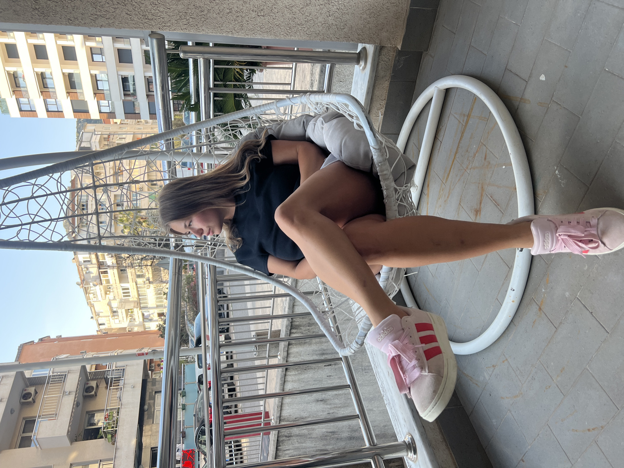 A young woman is sitting in a white hanging chair on a balcony overlooking a city. She is wearing a black t-shirt and pink Adidas sneakers. The balcony has a metal railing. The city behind the balcony is identified as Budva, Montenegro, based on the architecture and overall landscape. Several buildings with multiple stories are visible, along with vehicles parked along the street below. The day appears to be clear and sunny.